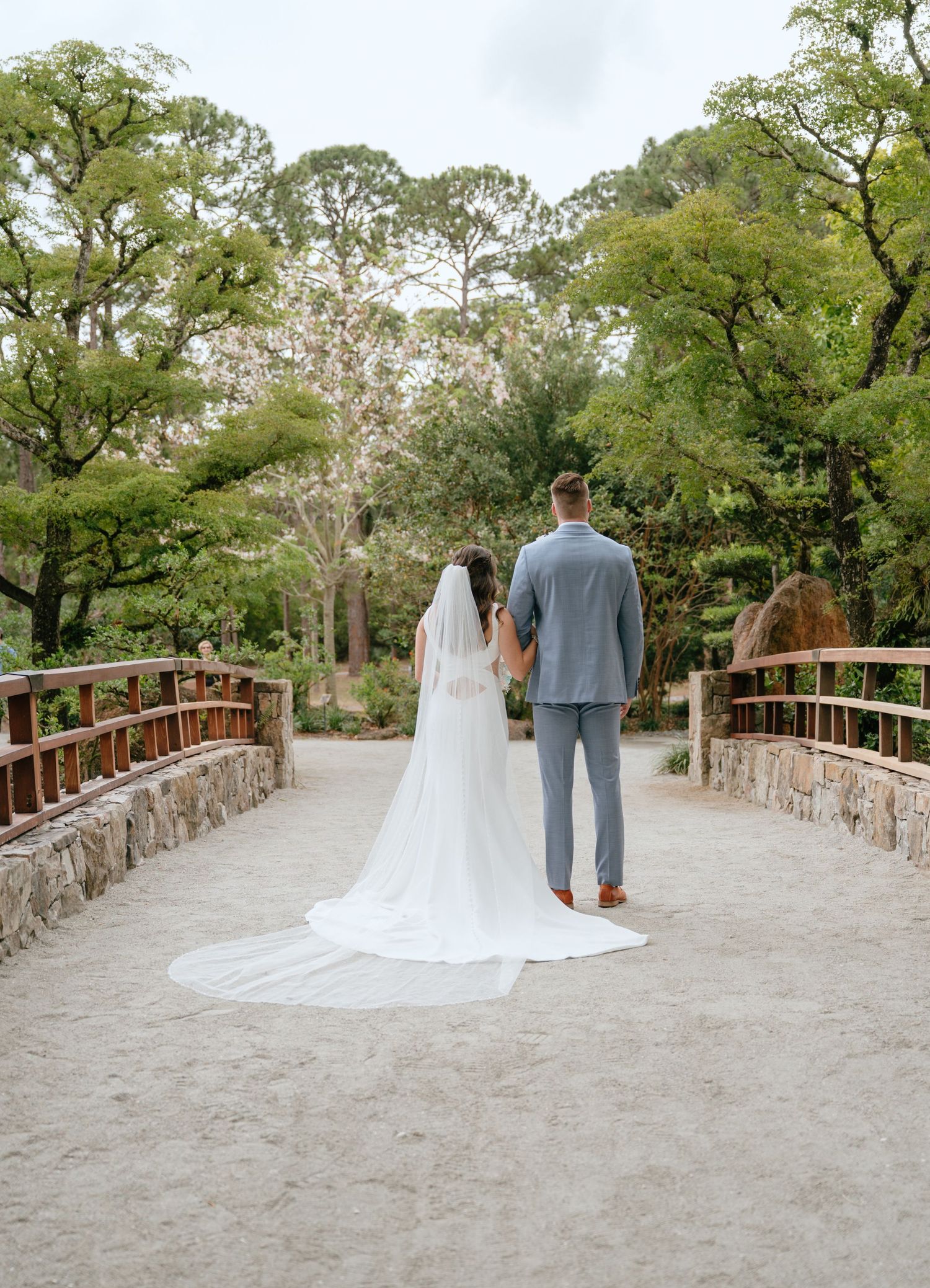Bride and groom hold hands on a stone bridge, her cathedral veil trailing behind in a lush garden.