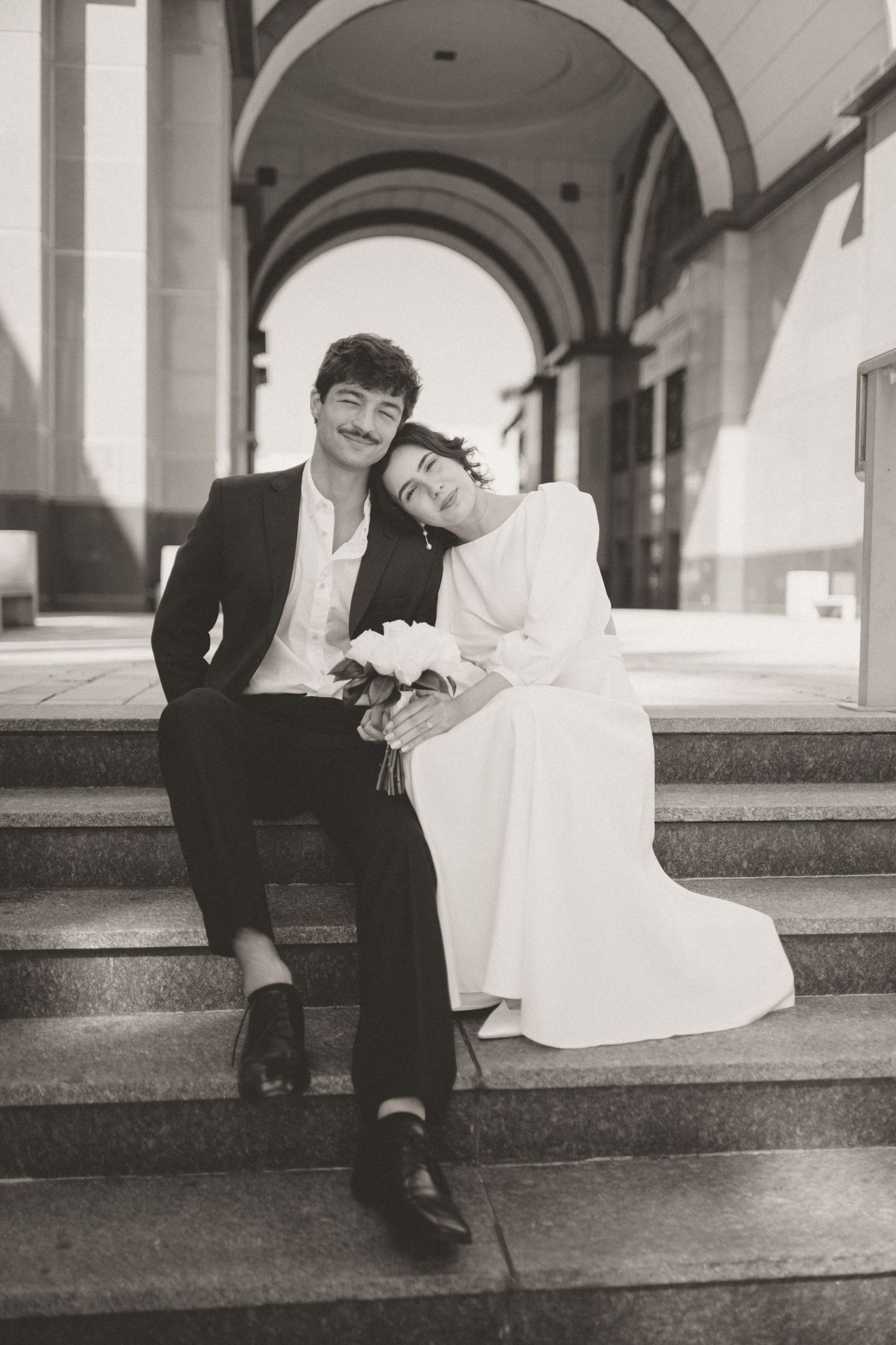 Black and white photo of newlyweds sitting on steps, bride resting head on grooms shoulder holding bouquet.