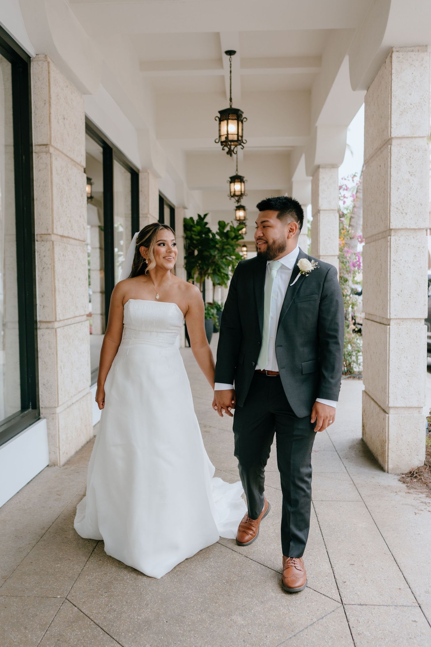 Newlyweds walk hand in hand along elegant covered walkway with hanging lanterns on wedding day.