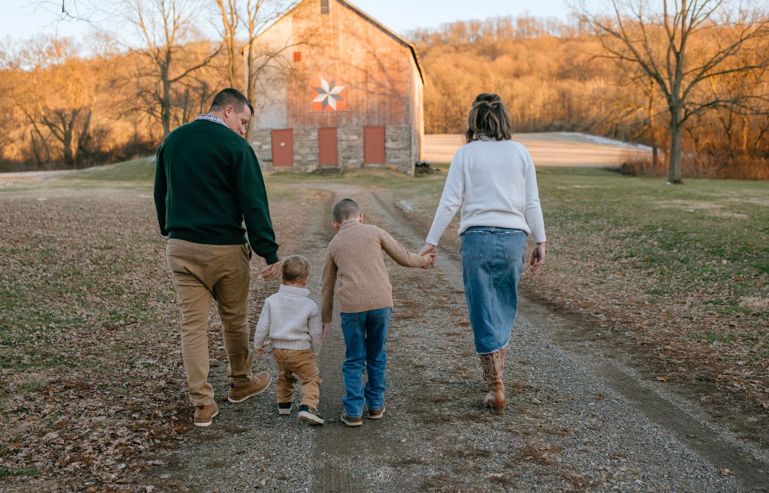 Family of four walking hand-in-hand down a gravel path toward a rustic red barn at golden hour.