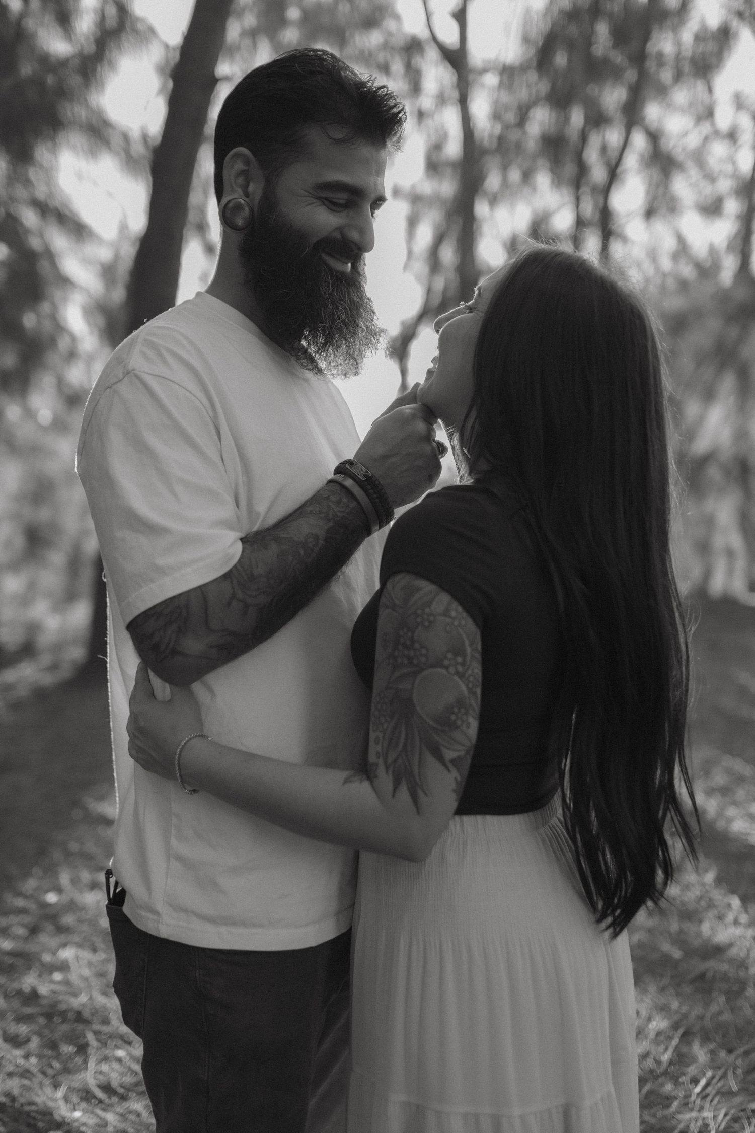 Black and white photo of tattooed couple sharing a tender moment in a forest.
