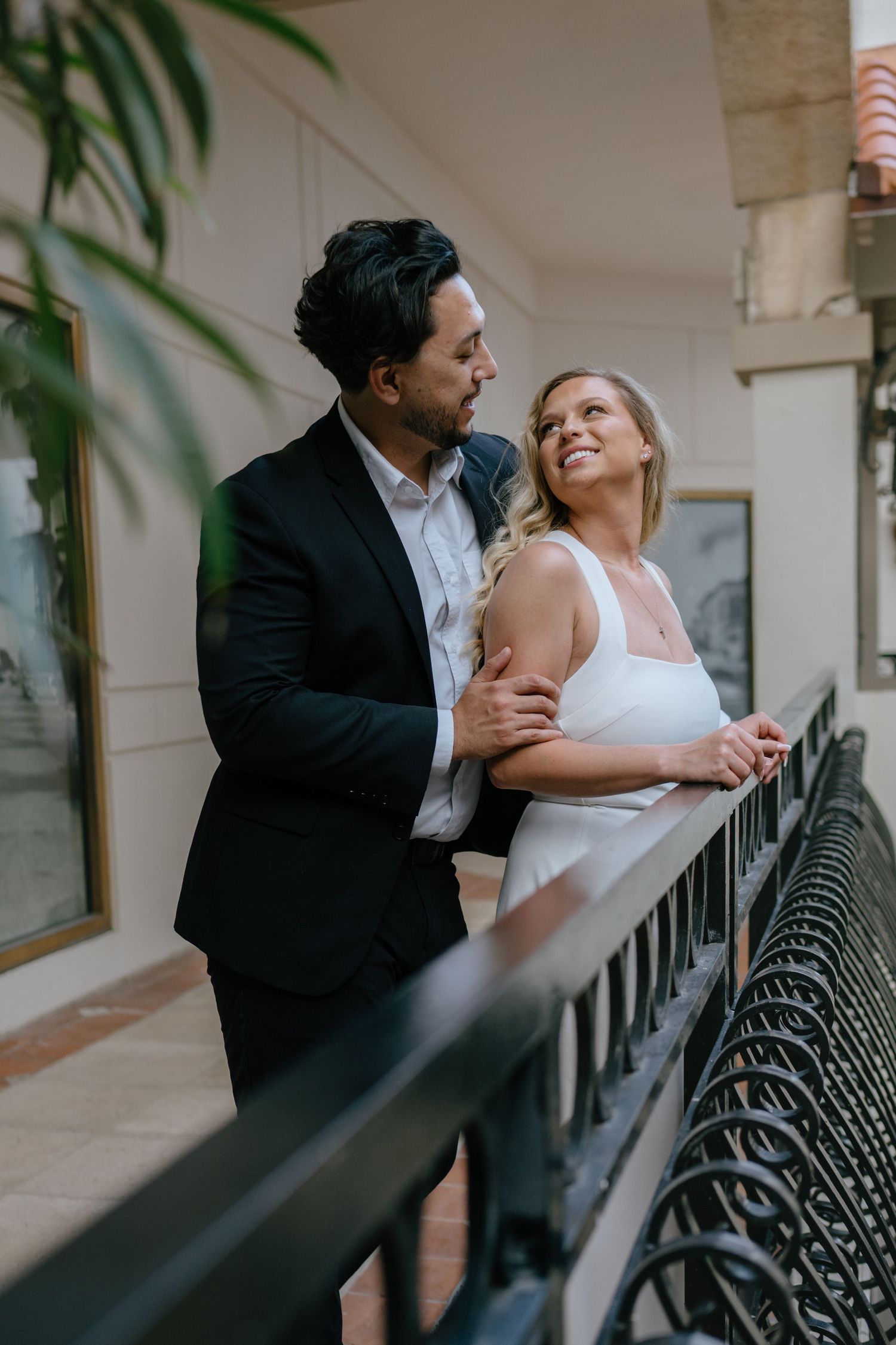 Couple in elegant wedding attire sharing a romantic moment on a balcony with iron railings.
