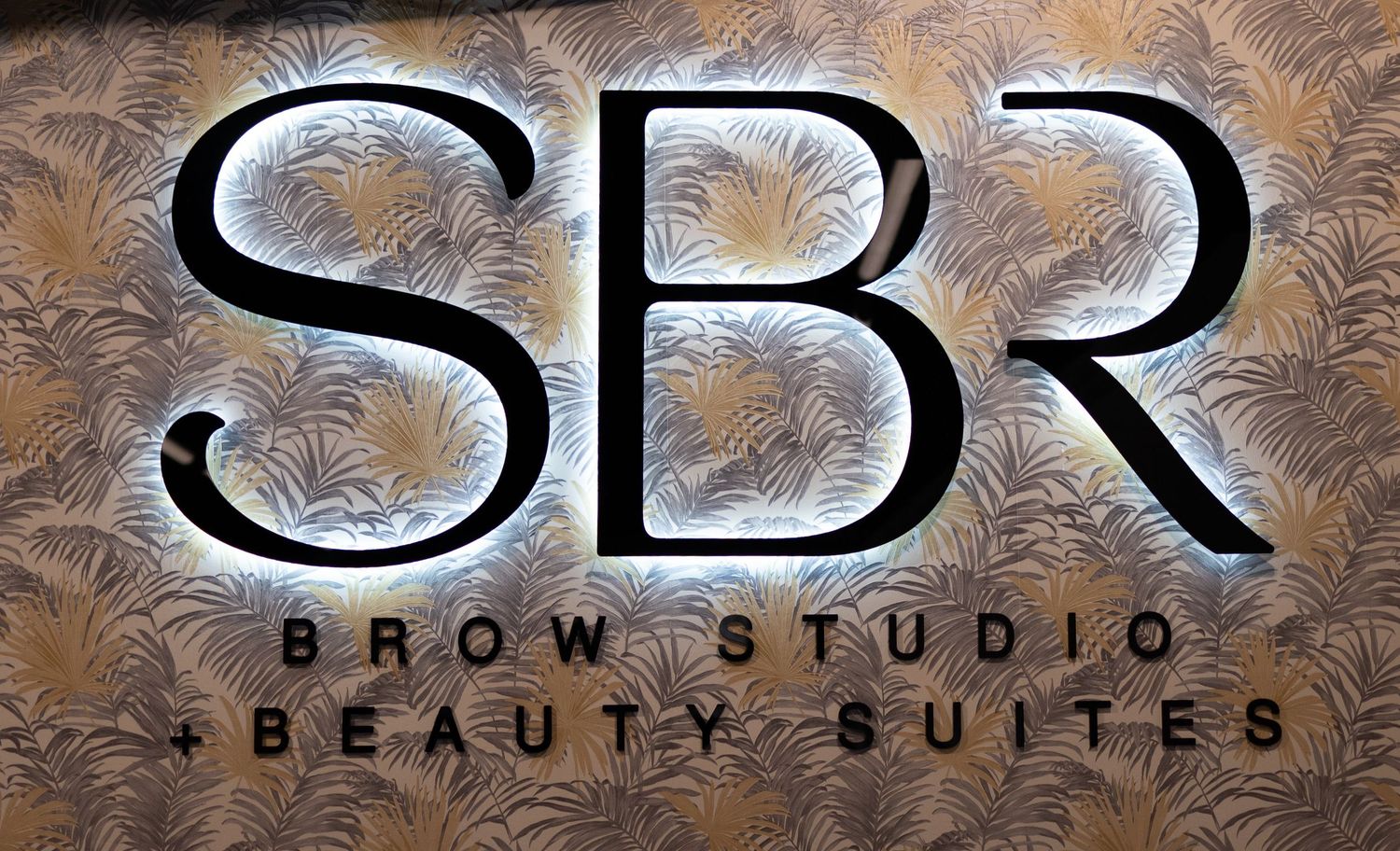 Illuminated SBR Brow Studio and Beauty Suites logo sign on decorative textured wall.