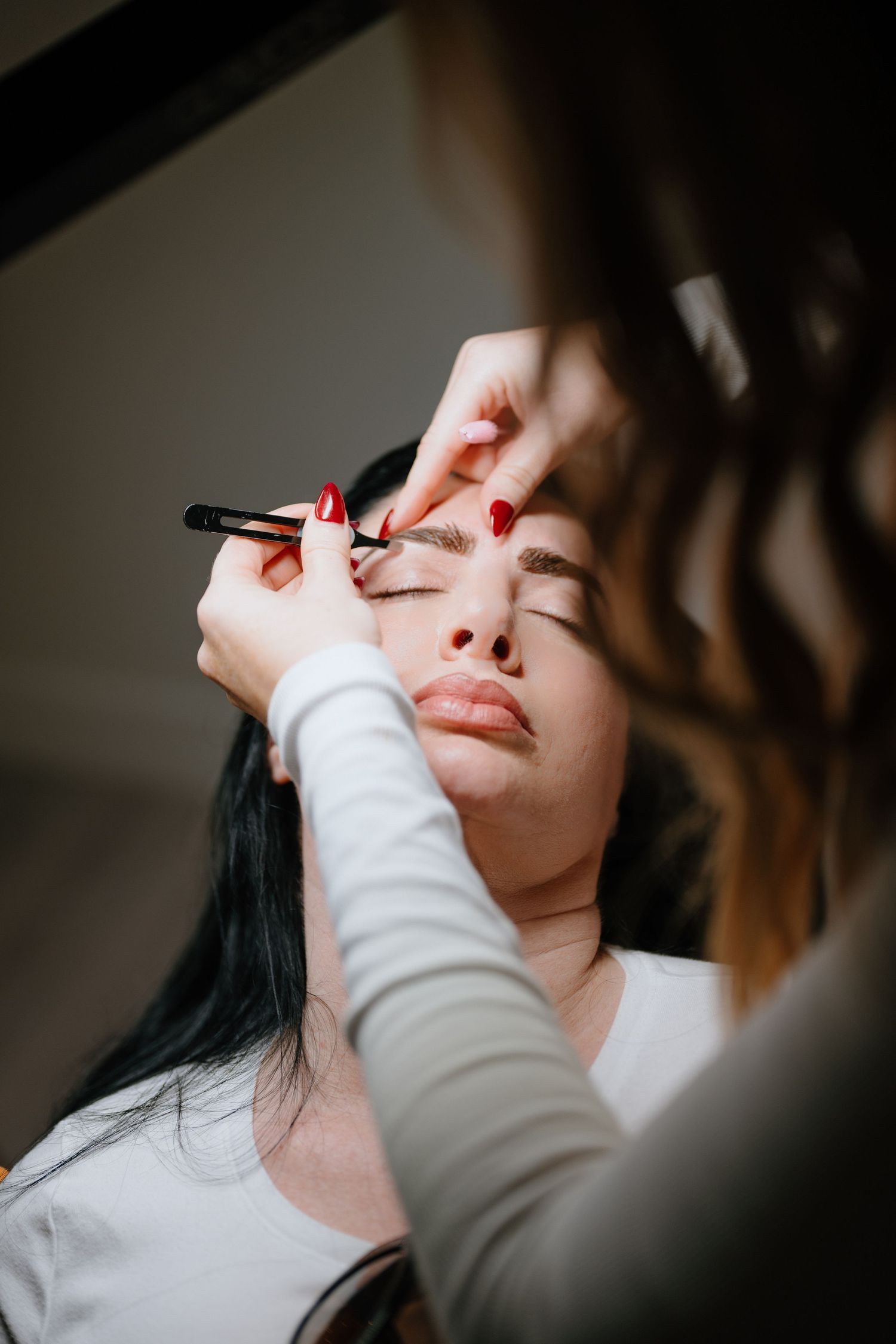 Beauty professional with red nails applies eyebrow makeup to a dark-haired female client using a precise brush.