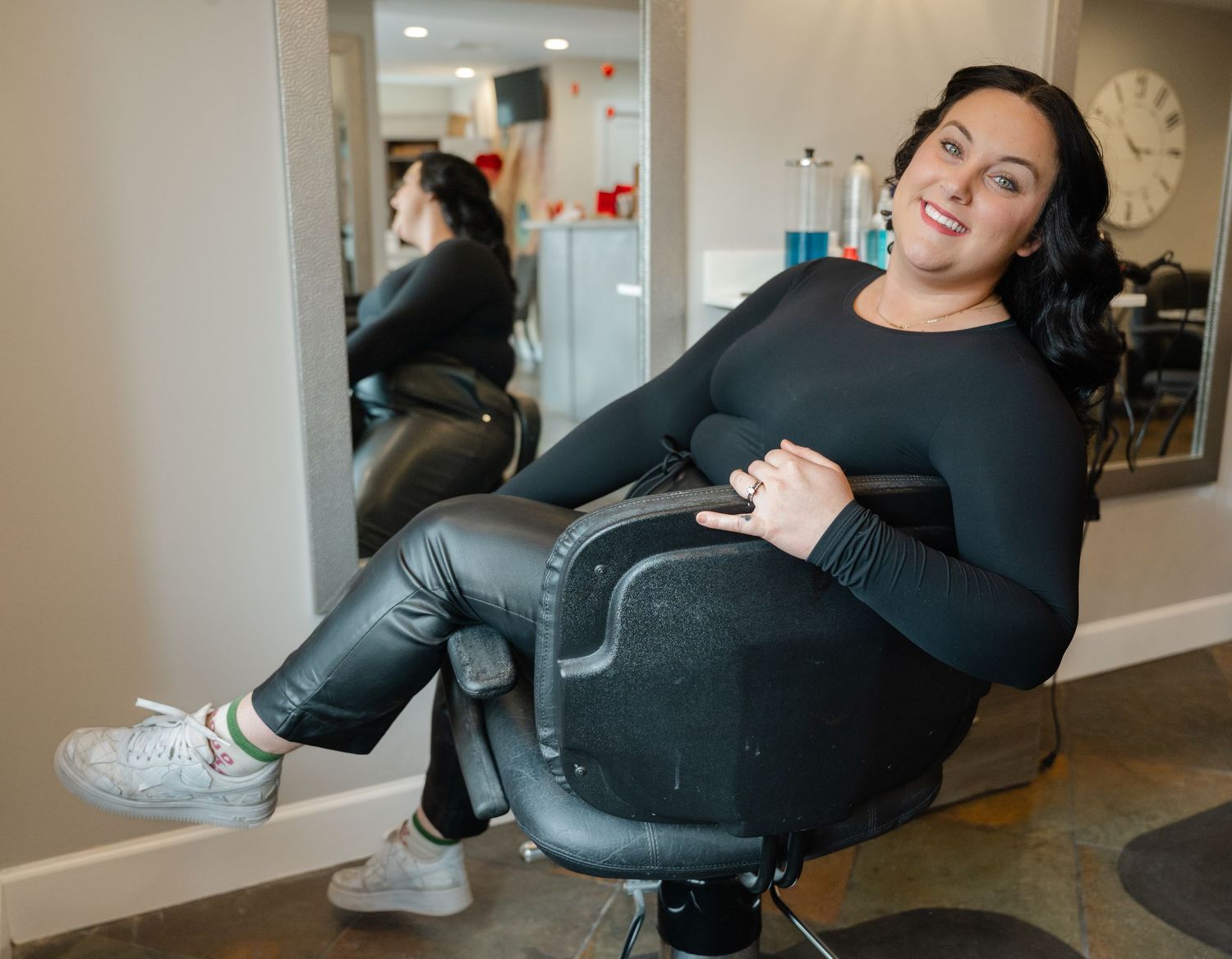 Woman in black outfit sitting in salon chair smiling, reflected in mirror behind her.
