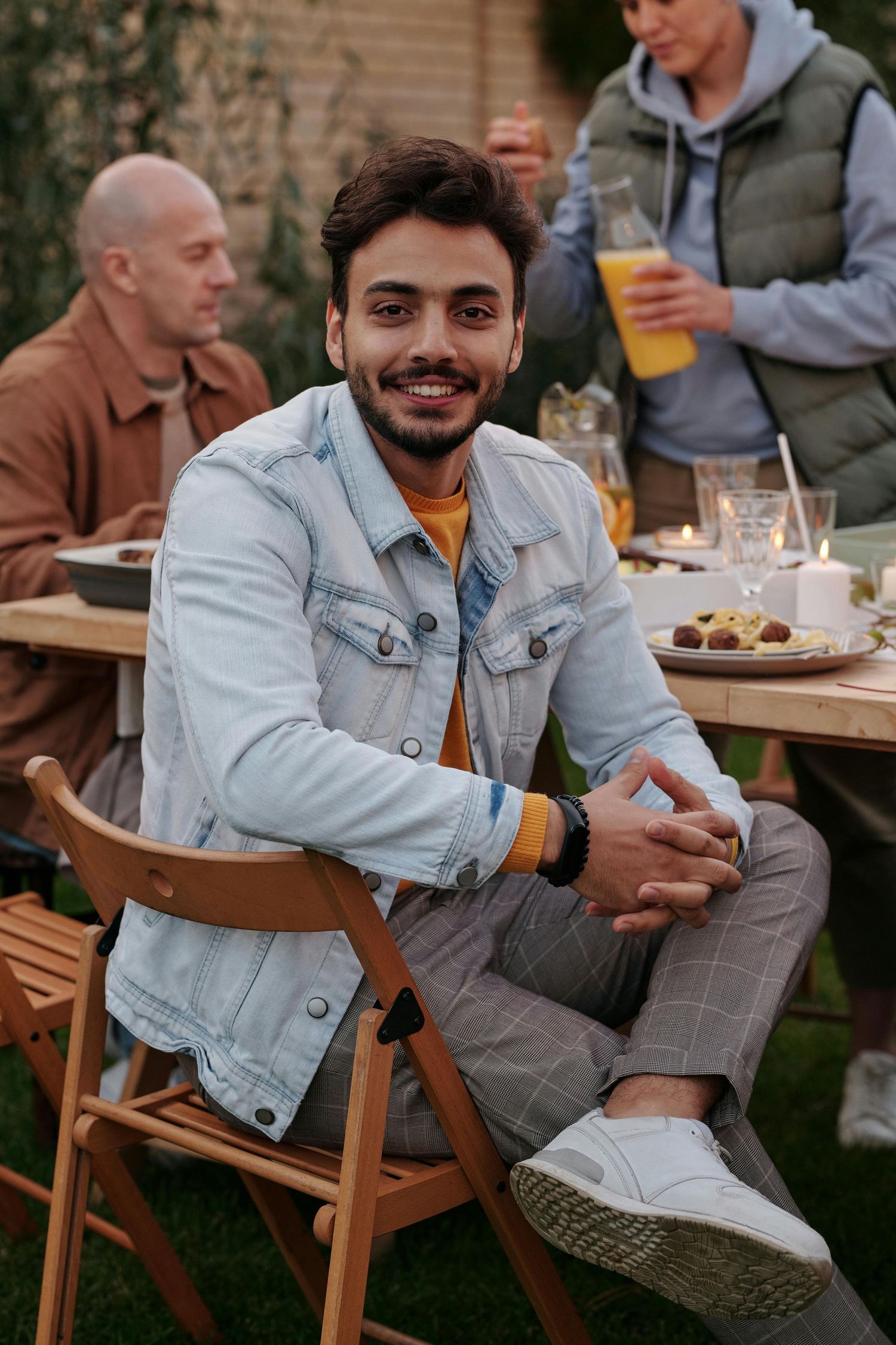 A person in a light denim jacket and orange shirt sits casually on a wooden chair at an outdoor gathering.