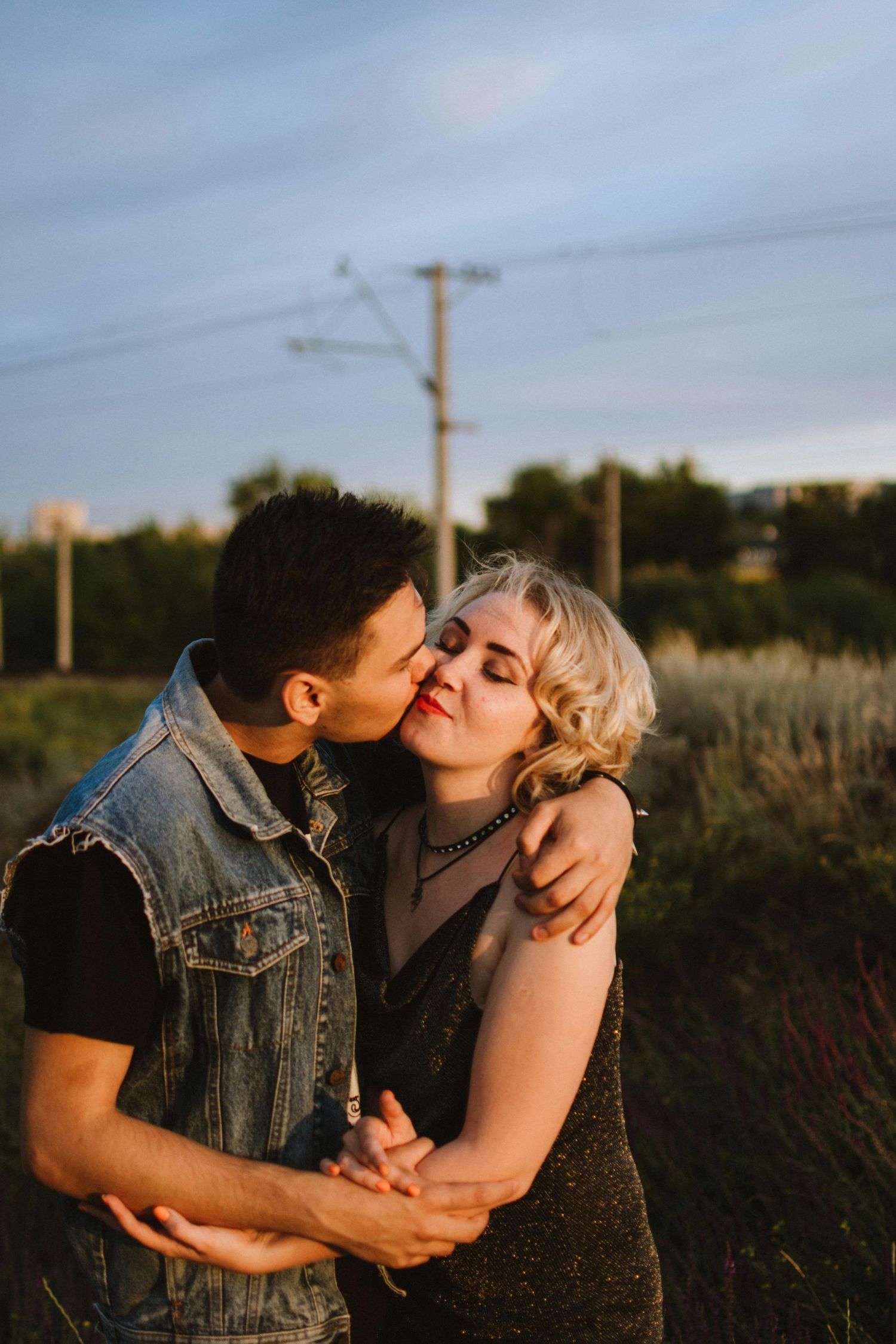 A couple embraces and kisses in a field at sunset with utility poles visible in the background. Top Photographer Scottsdale.