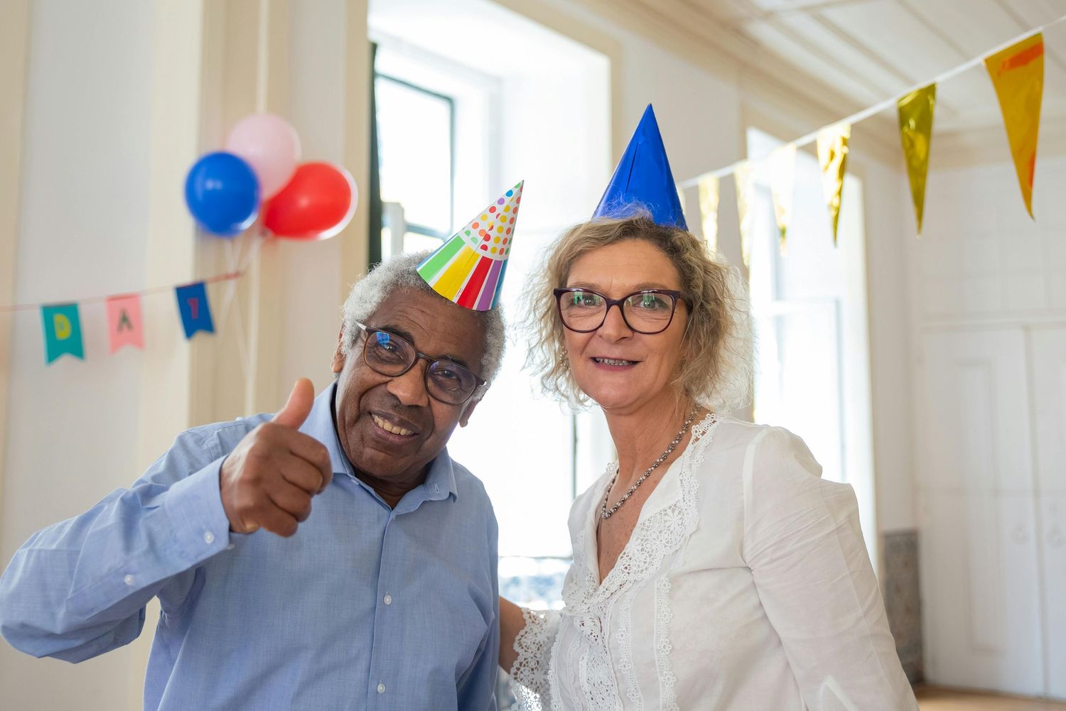 Two people wearing party hats celebrate together with balloons and festive bunting decorations in a bright room.