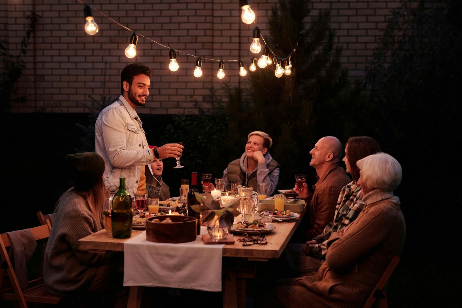 People enjoying an outdoor dinner party under string lights with a brick wall backdrop and cozy ambiance.