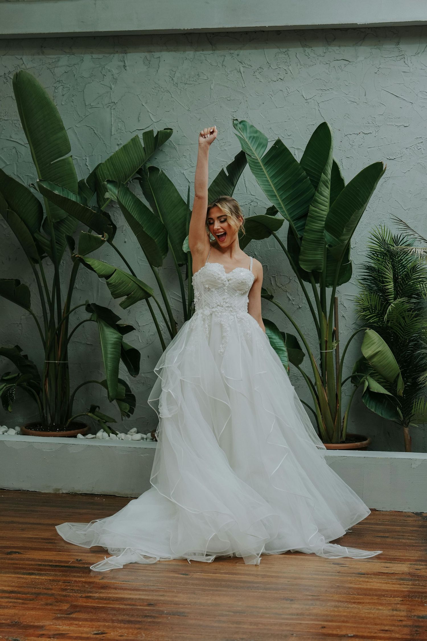 A bride in a flowing white gown poses joyfully against a backdrop of tropical plants and wooden floors. Leading Arizona Photographer.