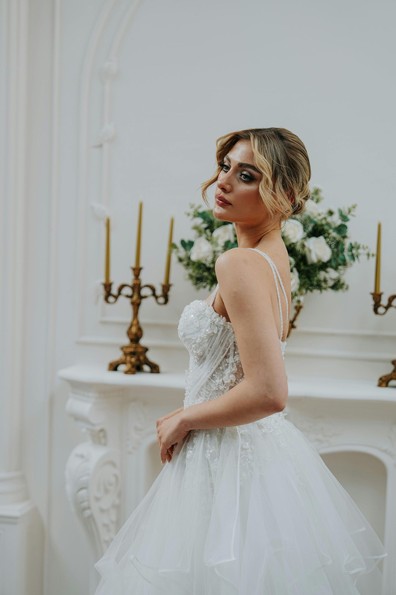 A bride in an elegant white gown poses dramatically near a classic white fireplace with gold candelabras. Leading Arizona Photographer.