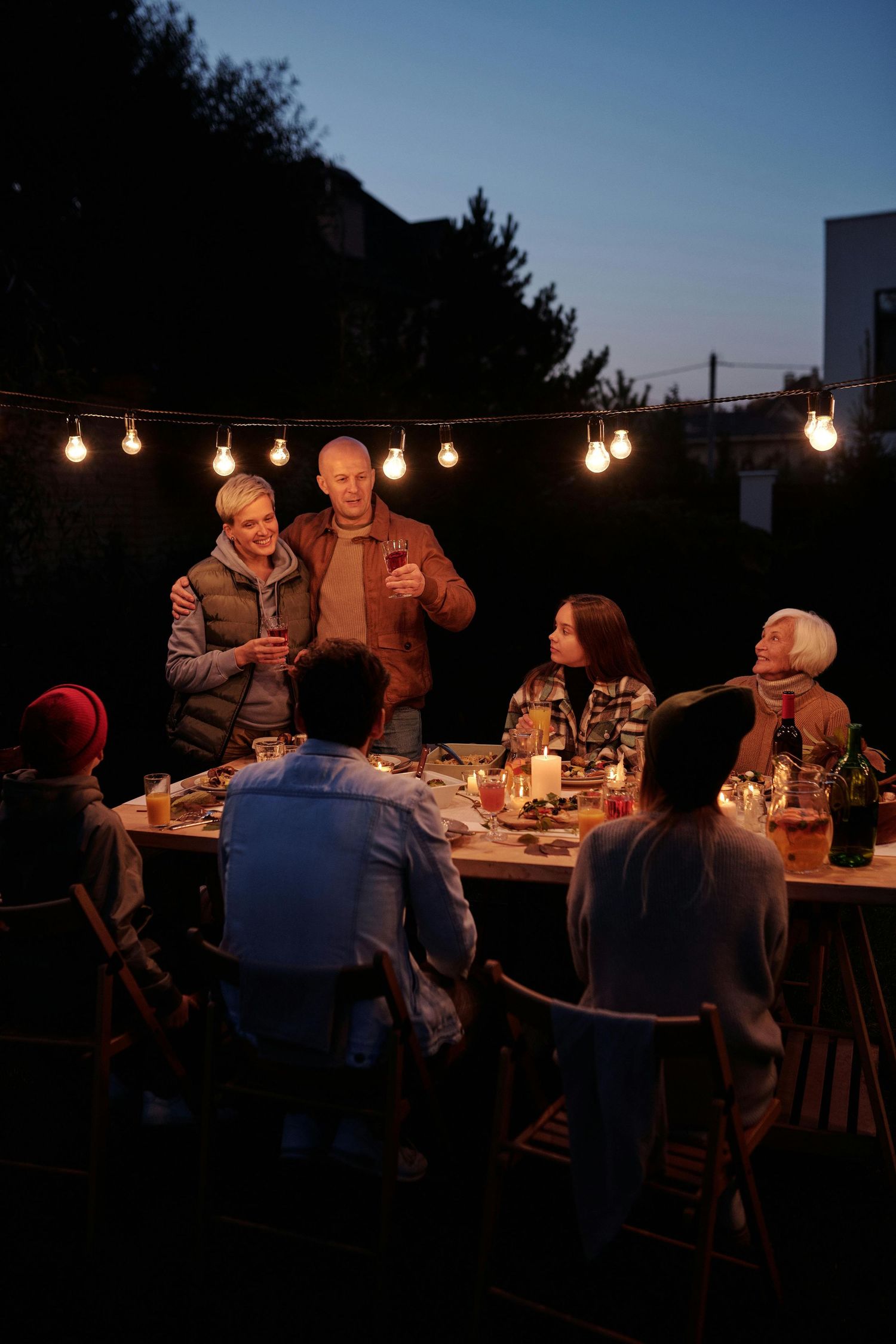 Evening outdoor dinner party with string lights creating warm ambiance as guests gather around a long table.
