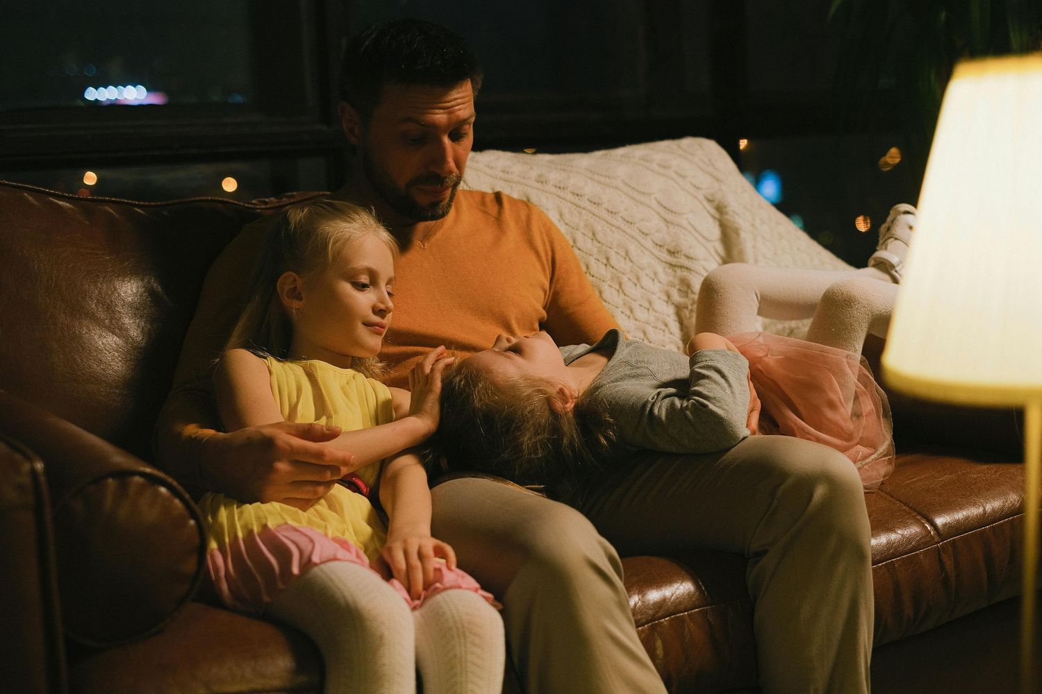 Two people relax together on a brown leather couch while watching something on a device in dim lighting.
