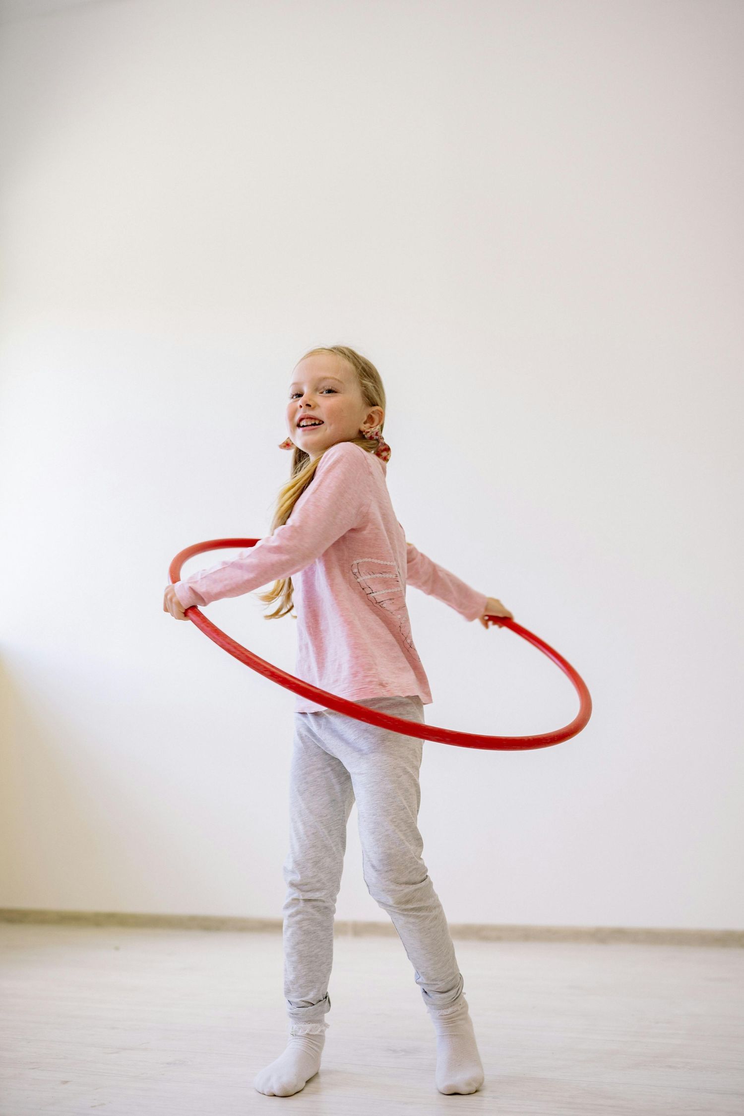 Arizona Daycare Photography Child in pink sweater and gray pants playing with a red hula hoop in a white room.