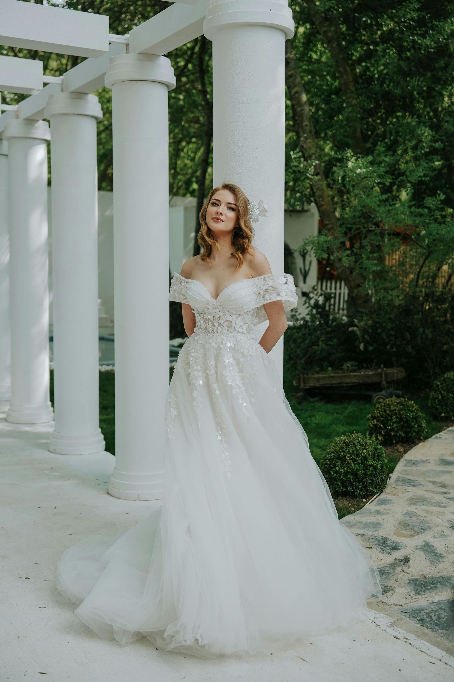 Arizona elopement. A bride in a flowing white off-shoulder gown poses elegantly among classical white columns in a garden setting.