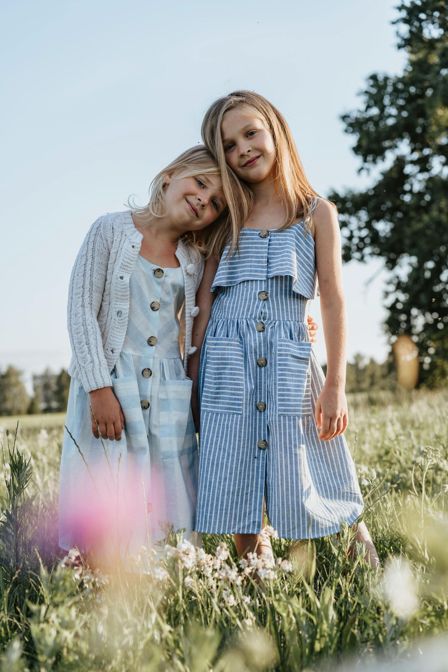 Two friends in summer dresses stand together in a sunlit meadow with wildflowers and trees in the background. Peoria Vistancia