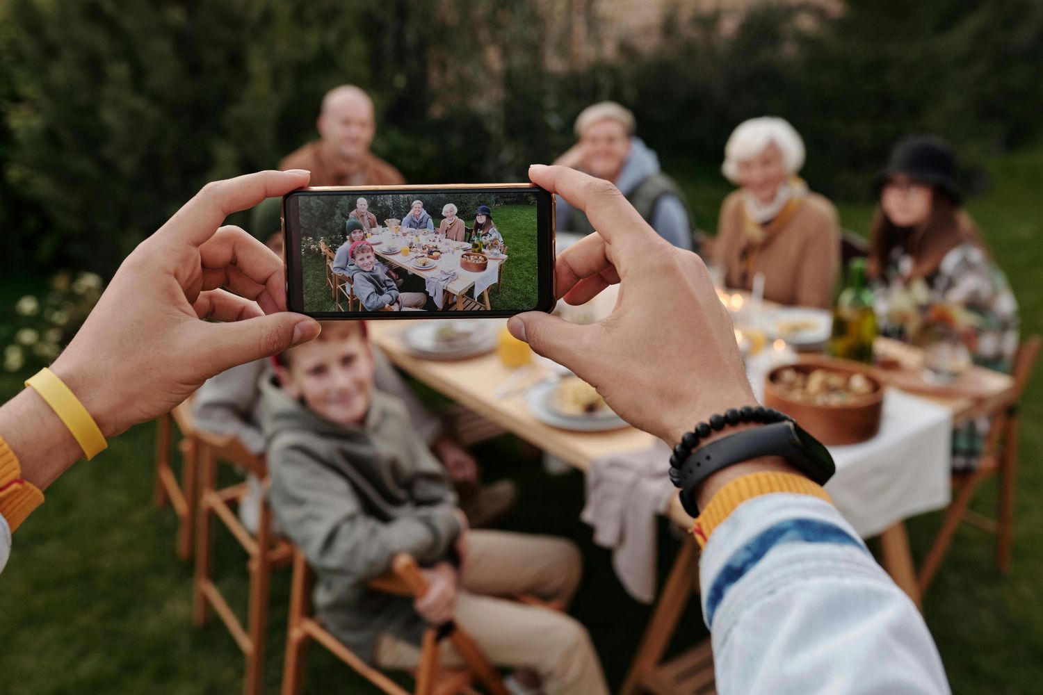 A group of people gather outdoors around a table for a casual summer meal in a garden setting.