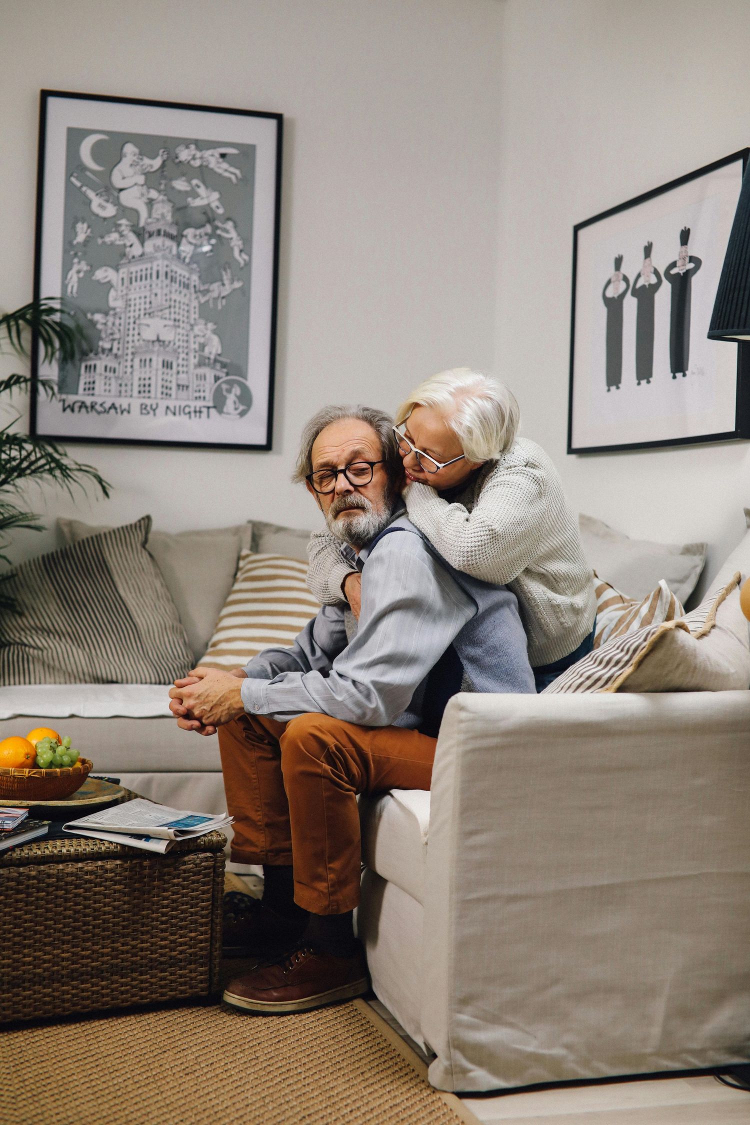 A cozy living room scene with two people sharing an intimate moment on a light gray sofa beneath framed wall art.