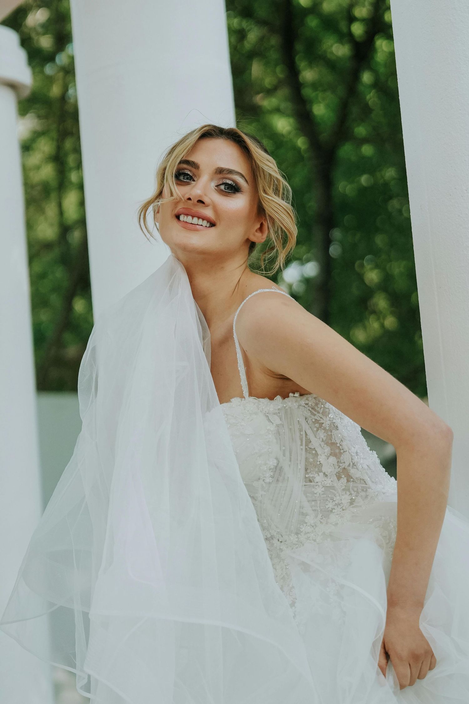 A bride in a white lace wedding dress and flowing veil poses outdoors against a green natural background. Top Photographer Scottsdale.