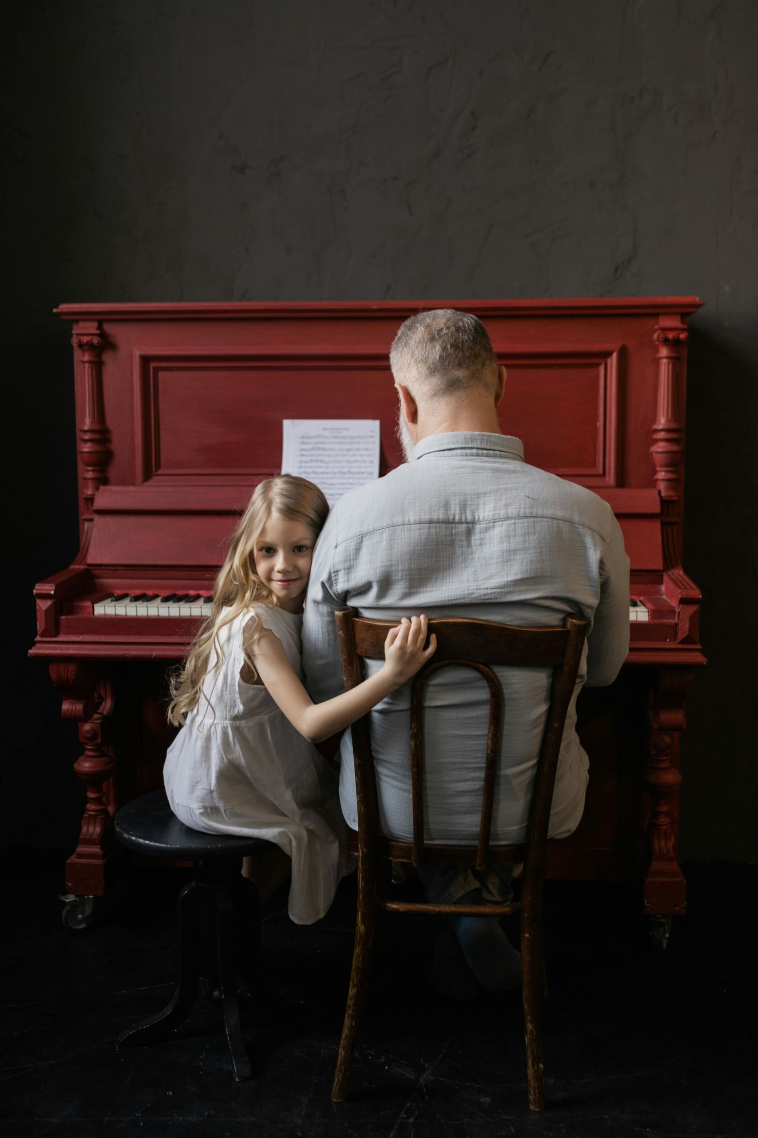 Two people sit together at a red upright piano against a dark background in a moody artistic series.