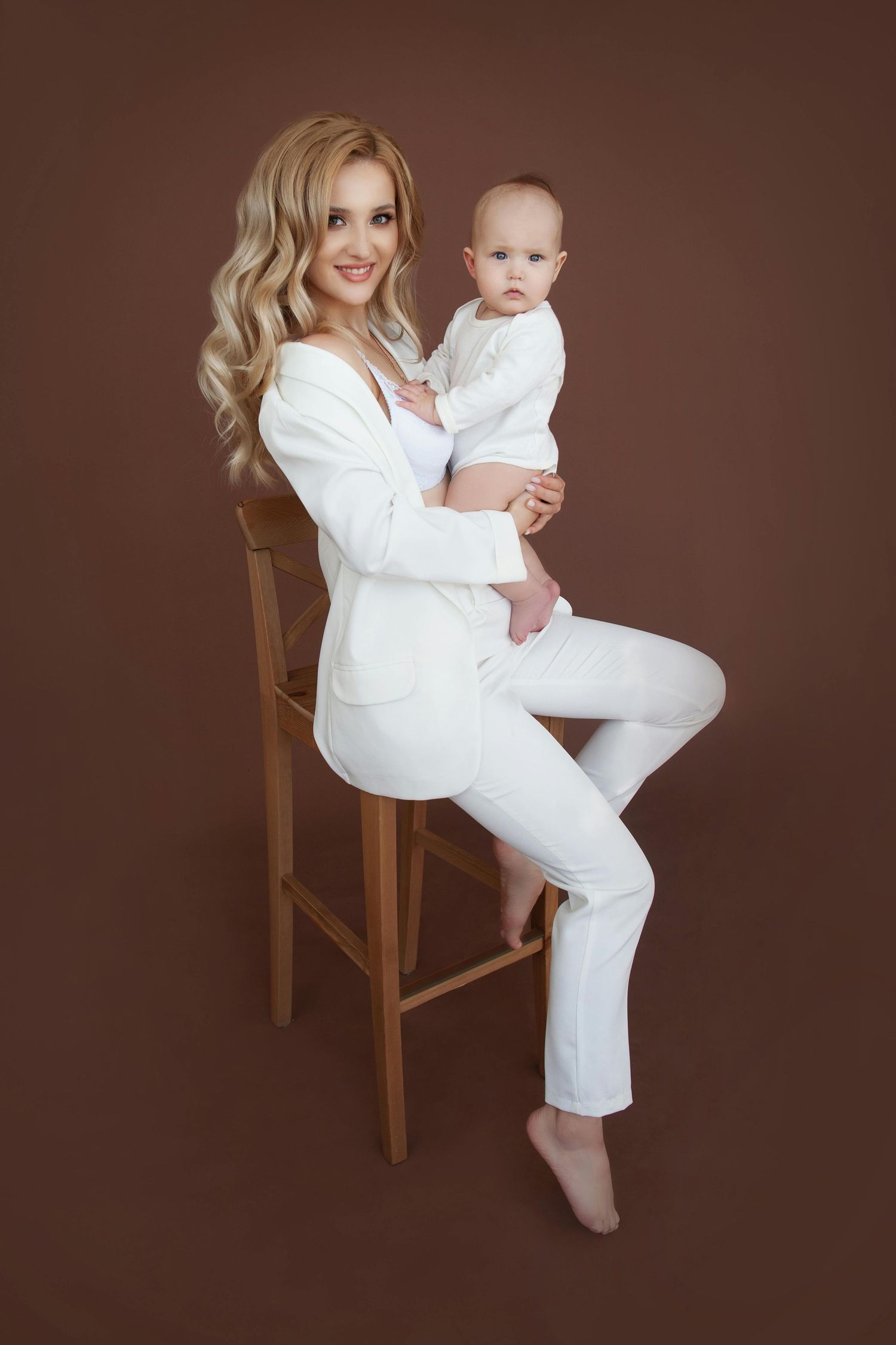 Studio portrait of a parent and baby in matching white outfits sitting on a wooden chair against brown backdrop. Desert Ridge Arizona.