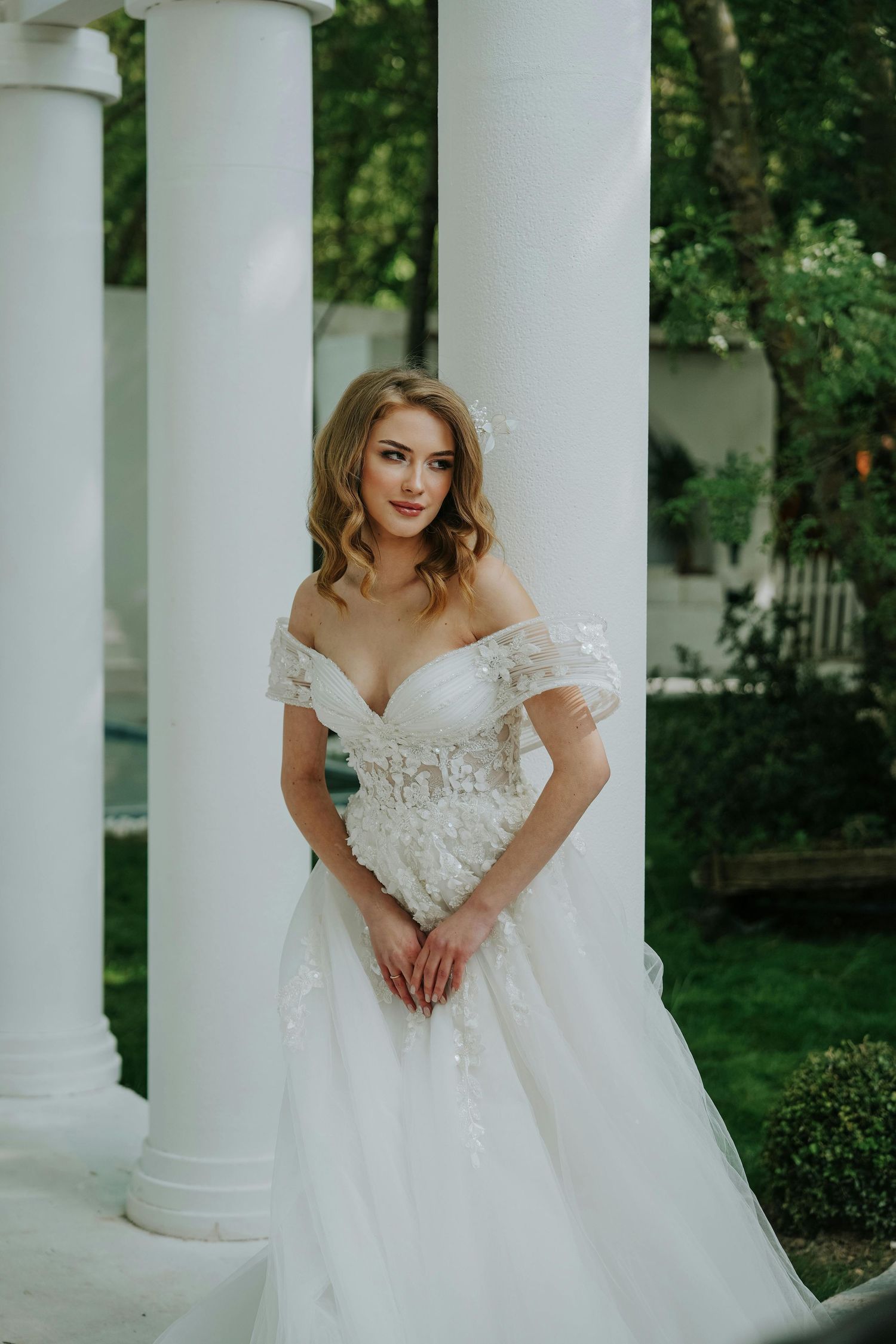 Beautiful bride in white lace off-shoulder wedding gown poses elegantly between classic white columns outdoors. Top Photographer Scottsdale.