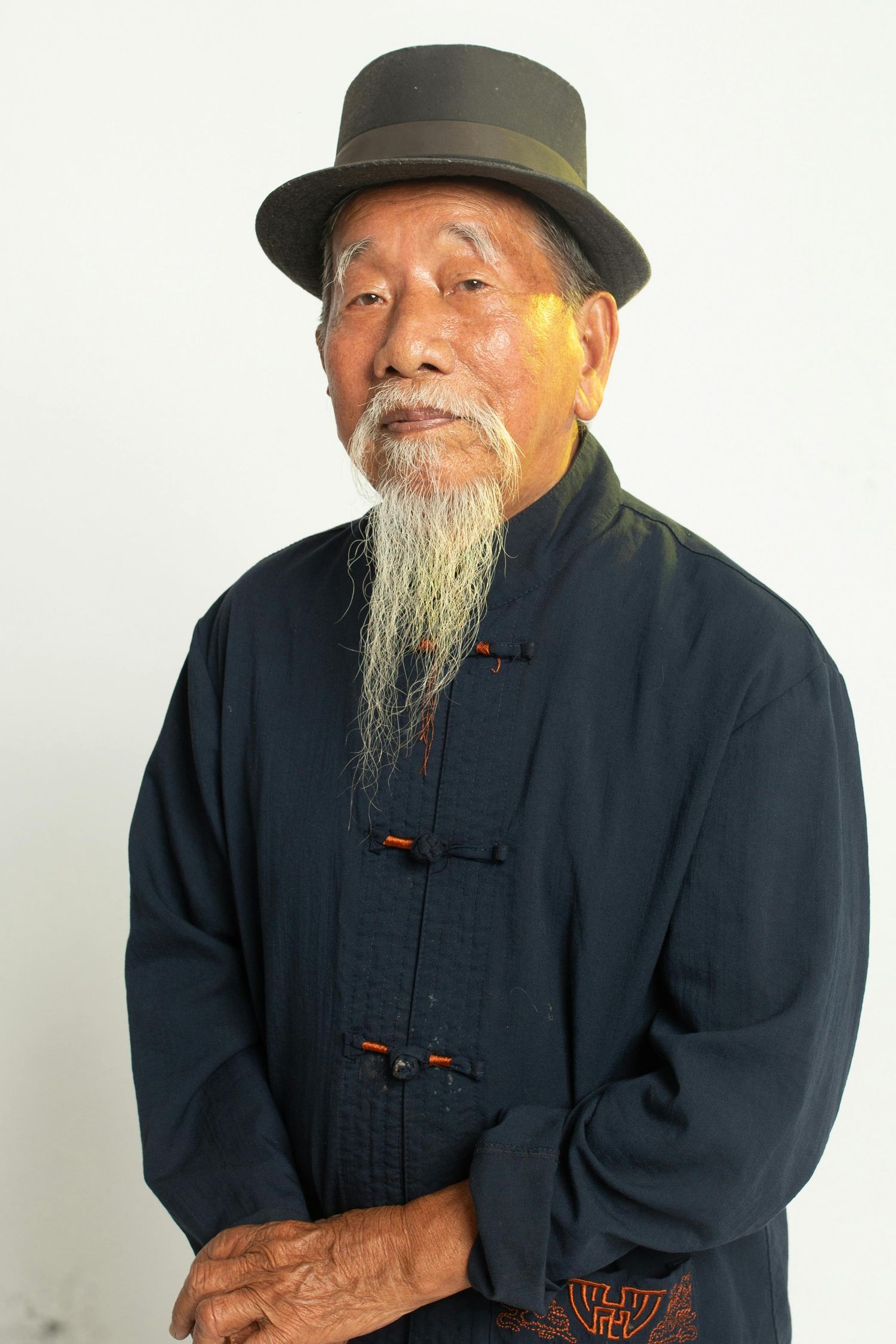 Elderly Asian man in traditional dark clothing and hat smiling warmly against white background.