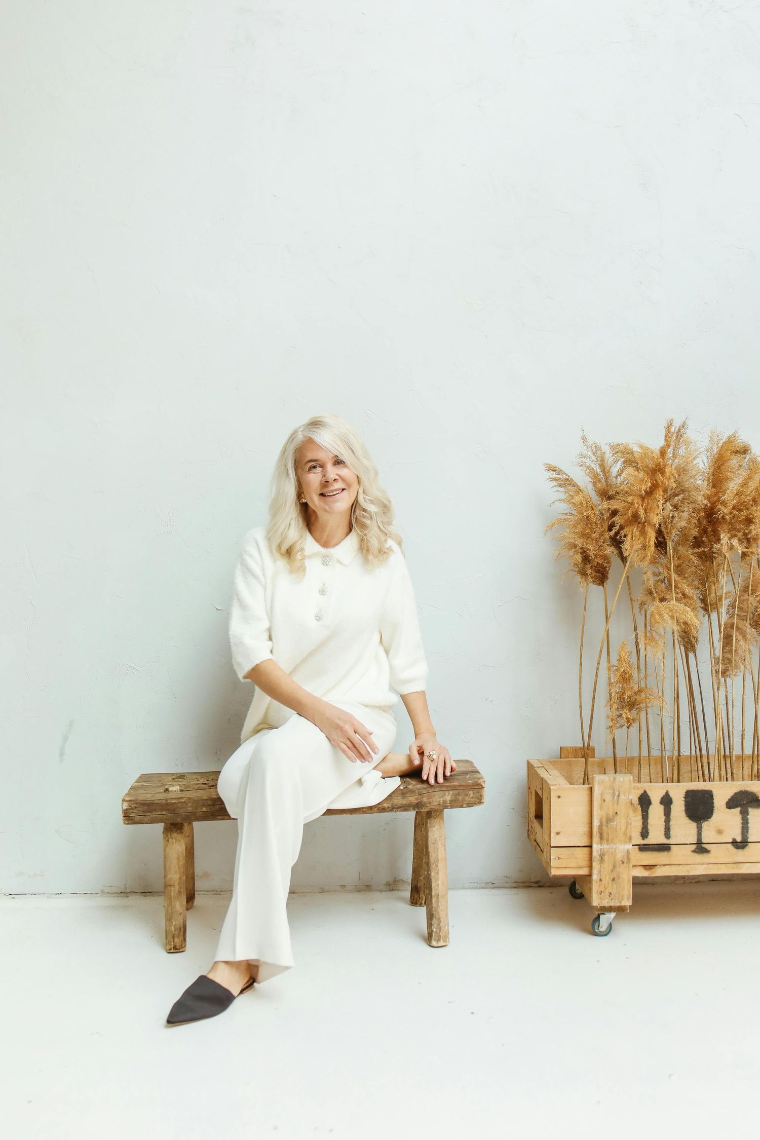 Person in white outfit sits on wooden bench beside decorative dried pampas grass arrangements against light wall.