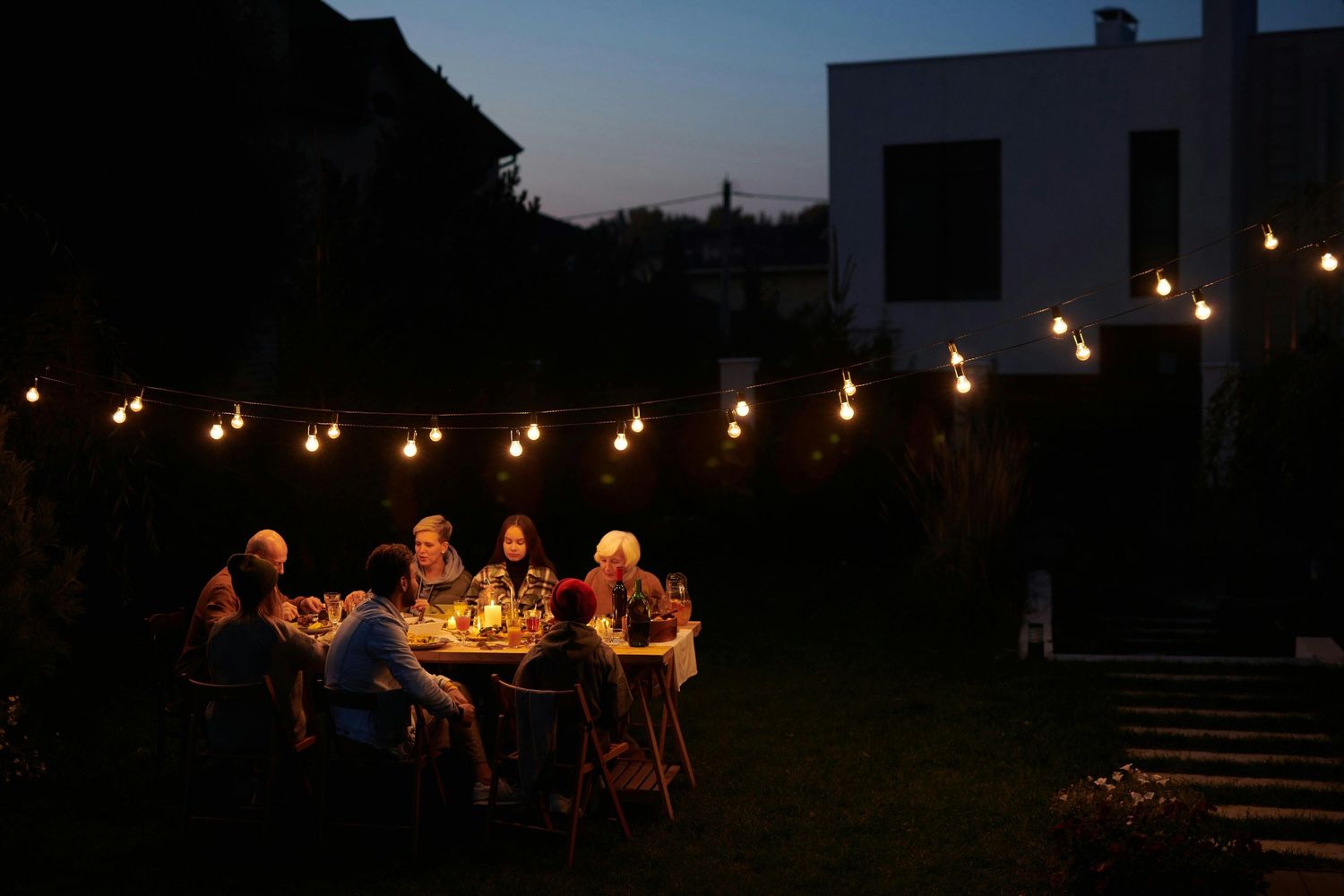 People gathered for an outdoor dinner party under string lights in a backyard at dusk.