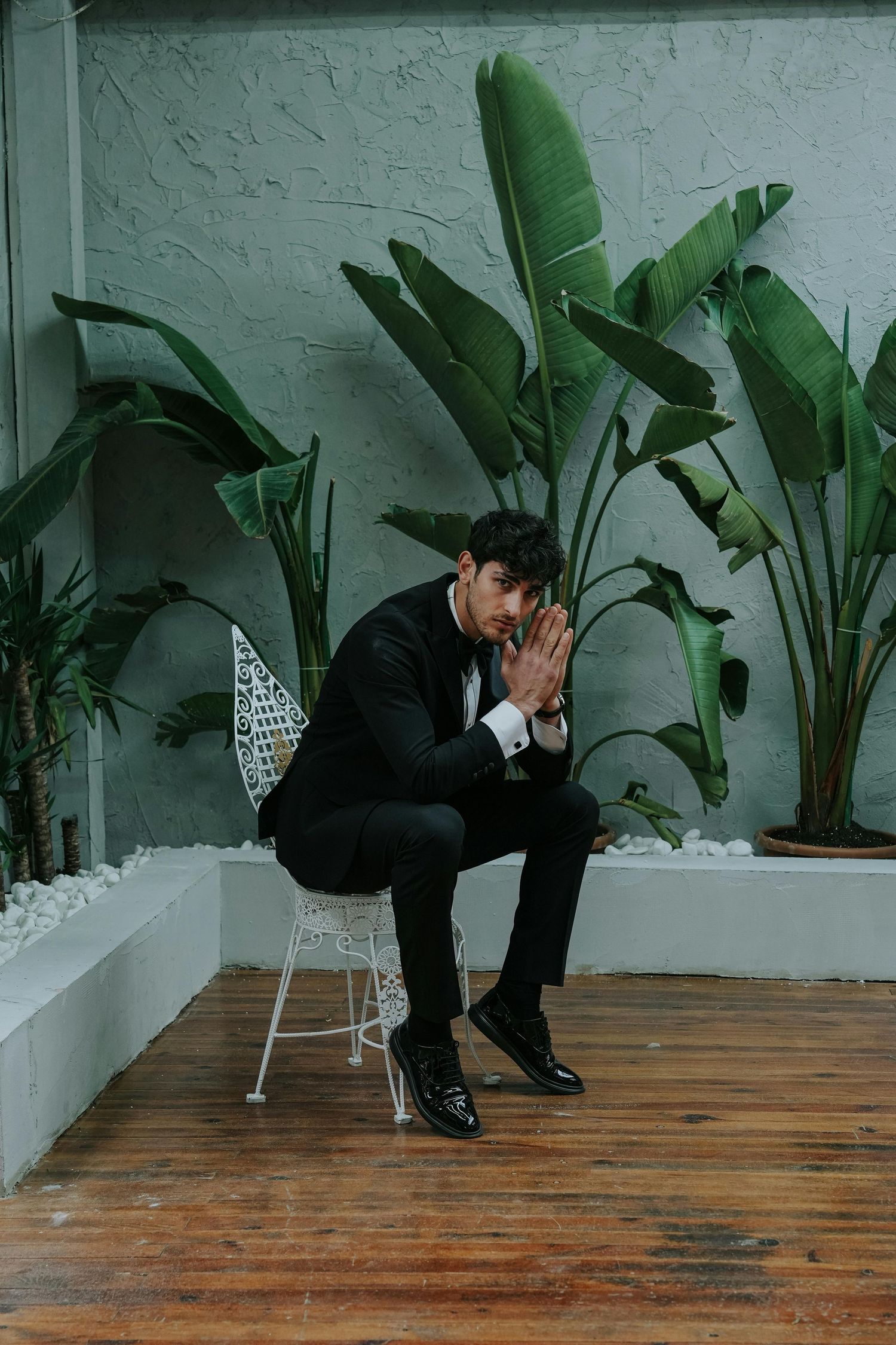Person in black suit sitting on wooden floor with tropical plants and white chair in the background.  Award Award-winning photographer.