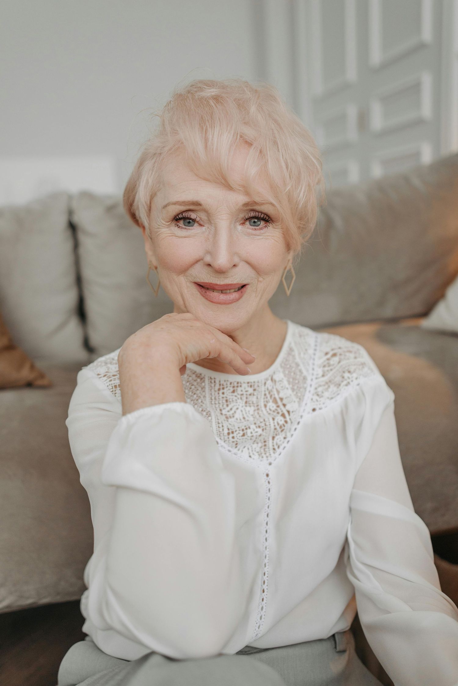 Elegant mature woman in white lace blouse smiles warmly while sitting on gray couch in soft lighting.