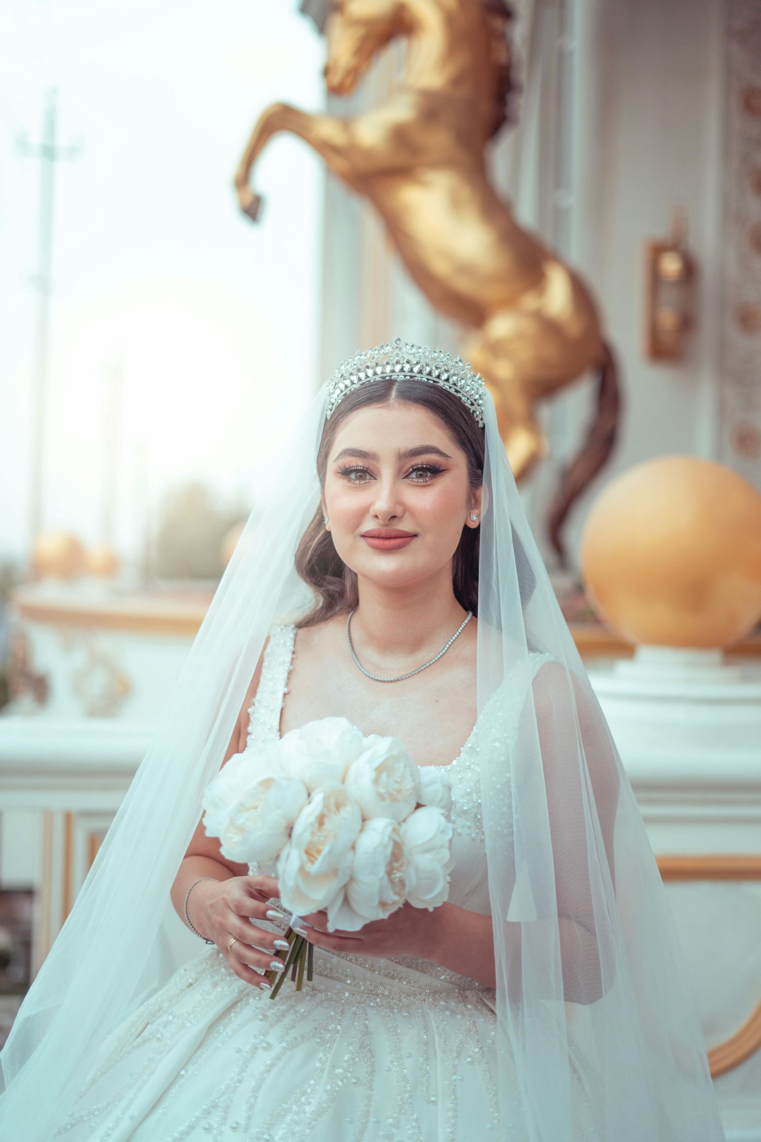 Scottsdale Arizona Photography Bride in a white lace gown with tiara and veil holds white bouquet with golden horse statue in background.