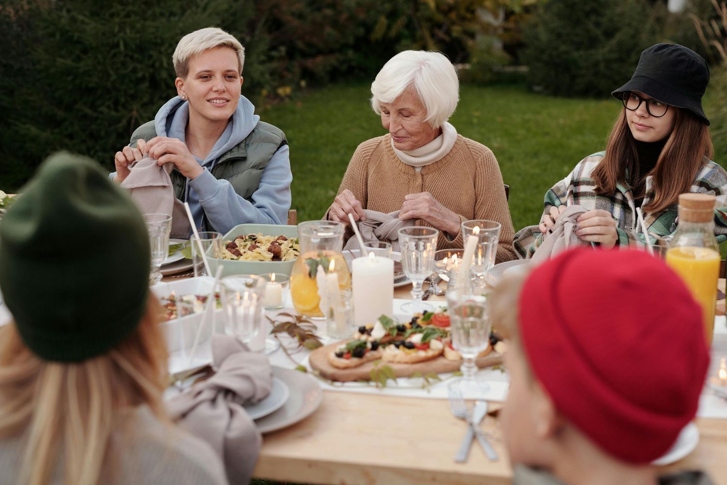 A group of friends enjoying an outdoor meal together at a wooden table with drinks and food spread out.