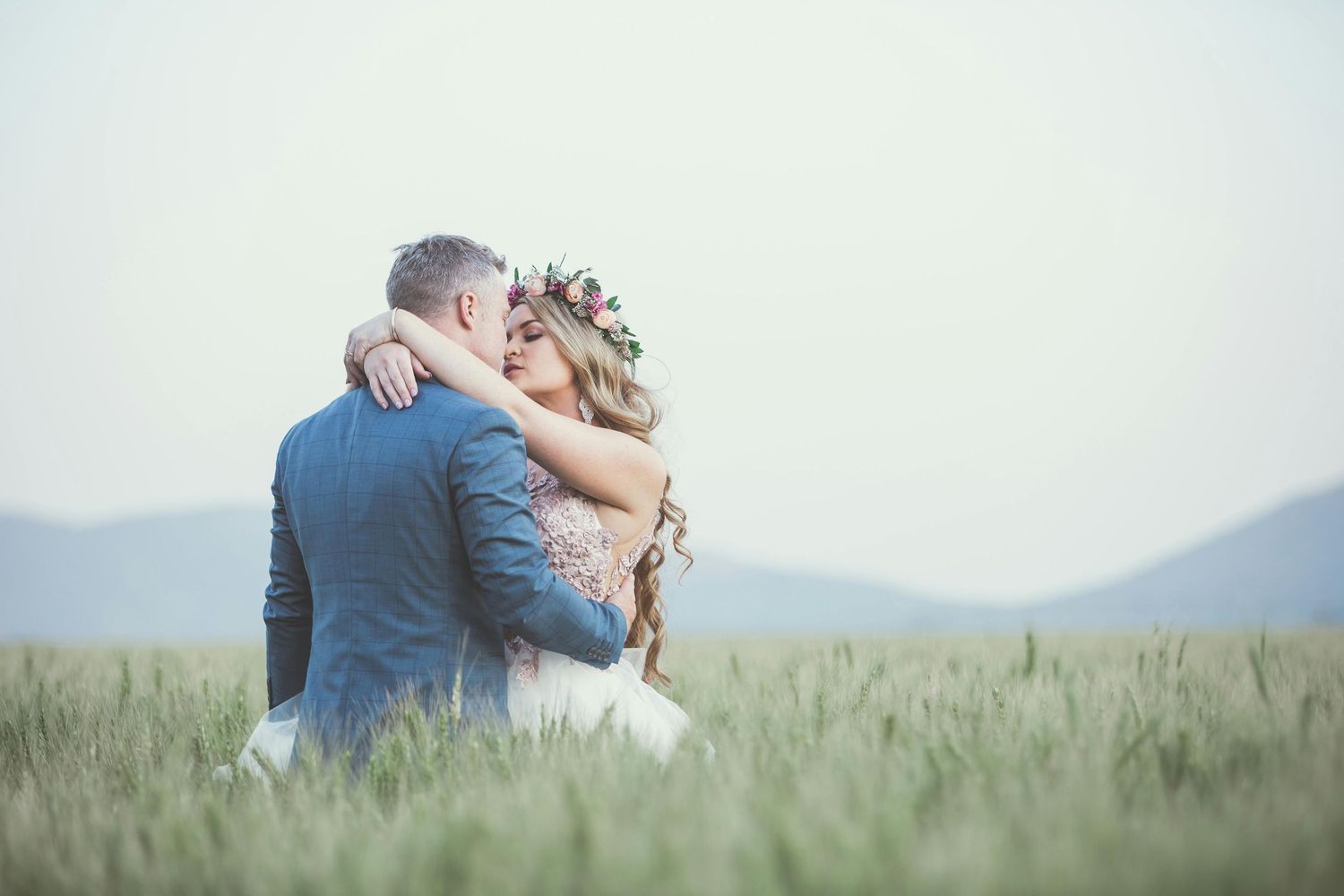 Two people embrace lovingly in a scenic green meadow with mountains visible in the distance.