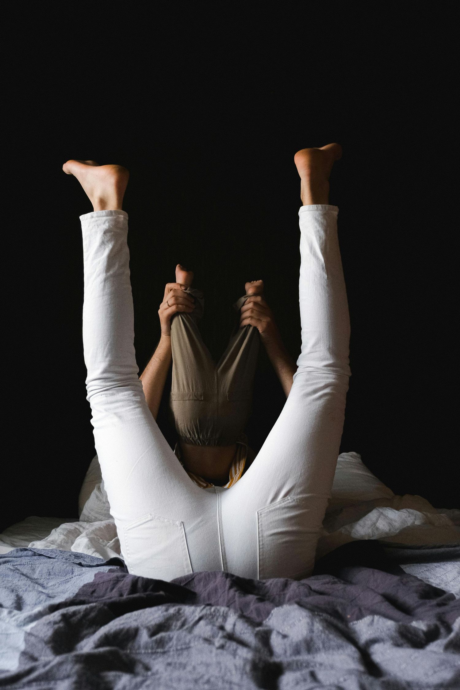 Home Photography Phoenix. Person wearing white pants lies on gray bedding while holding onto fabric in dramatic dark lighting composition.