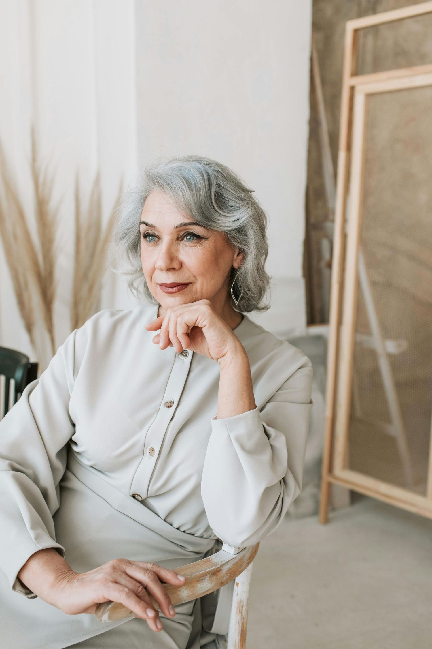 Elegant figure in a light beige outfit poses thoughtfully in an artistic setting with framed artwork.