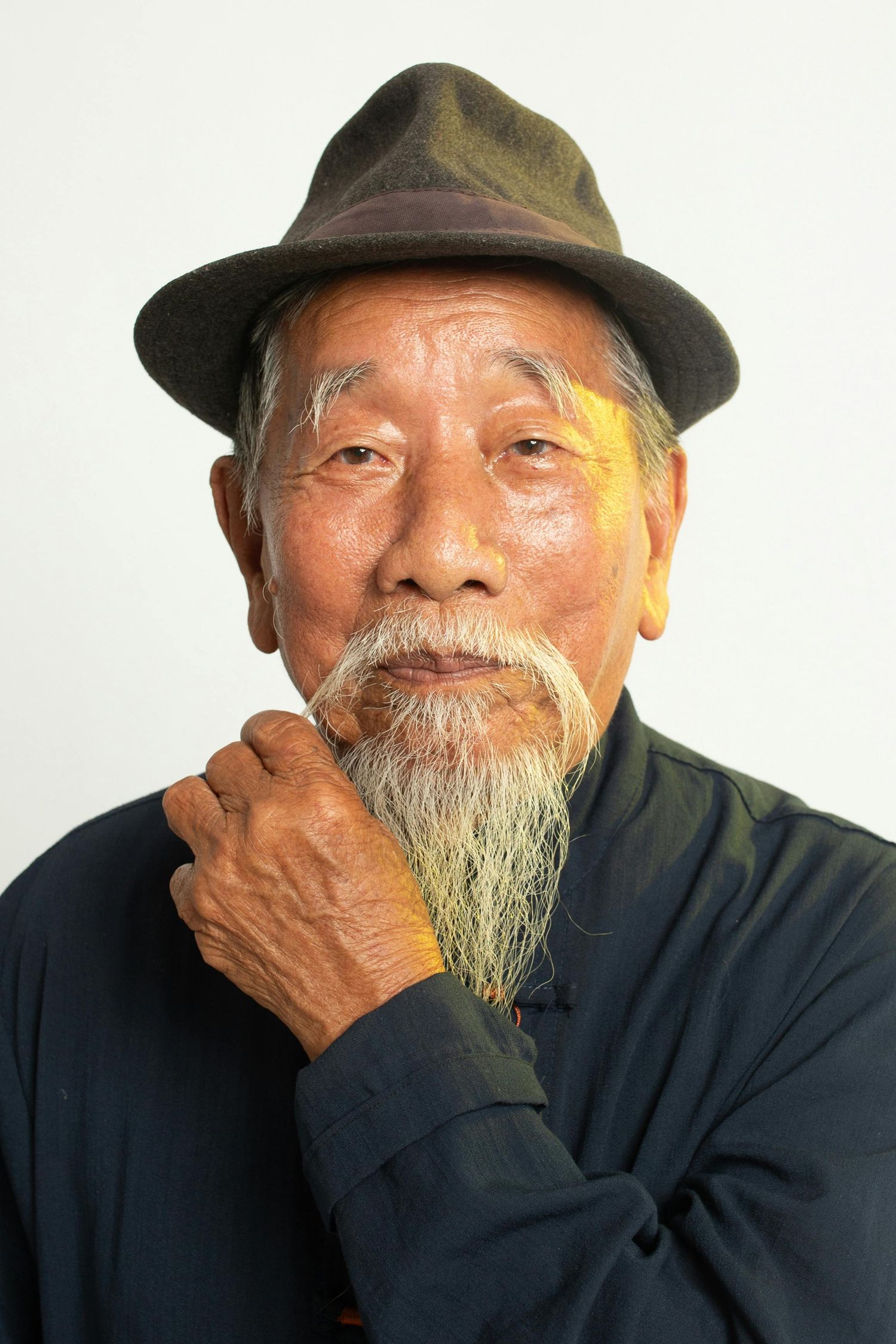 Elderly Asian man in dark attire covers mouth while expressing joy through his eyes.