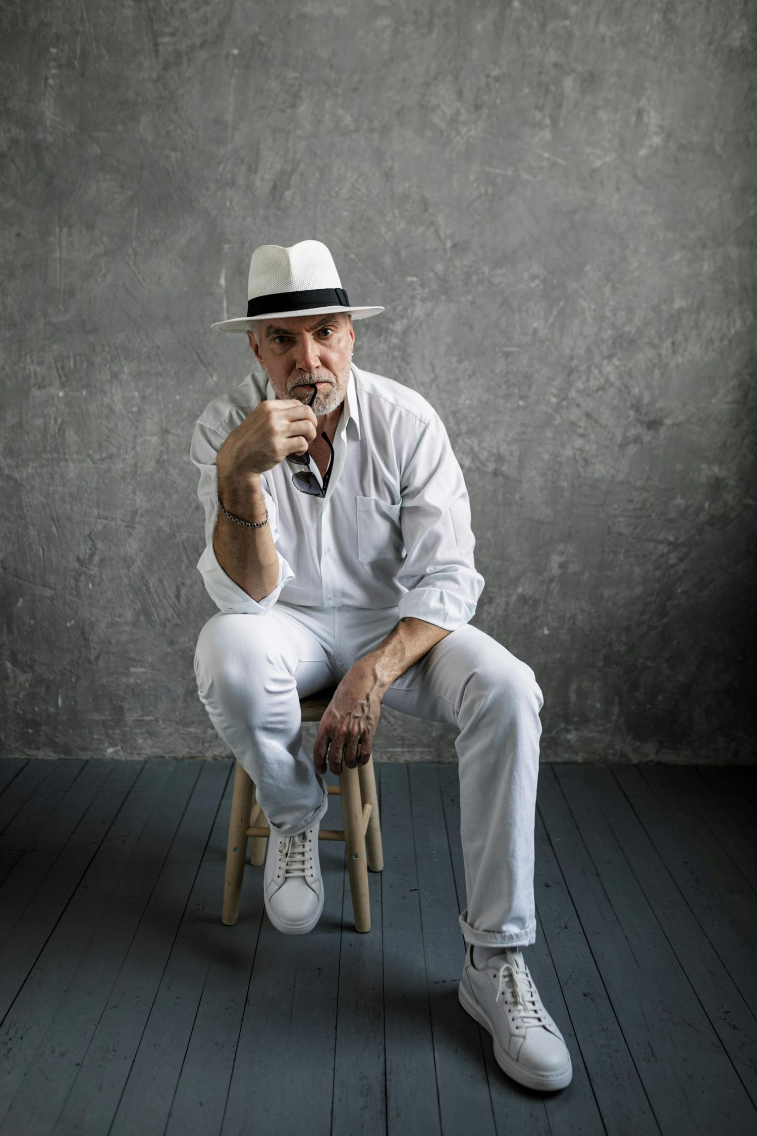 Paradise Valley Arizona Seniors Photography. Figure in white ensemble and fedora sits thoughtfully on wooden chair against textured wall.