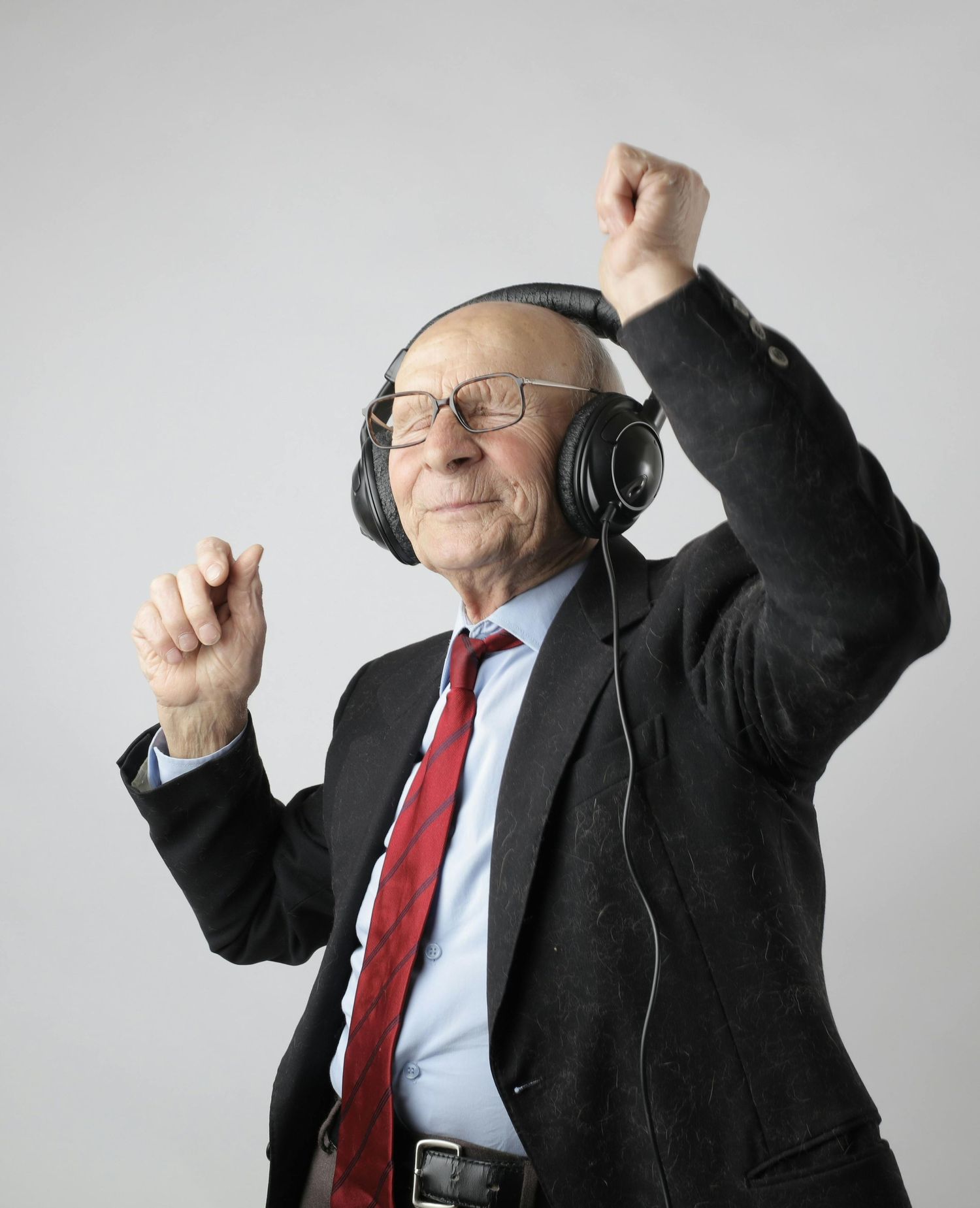 Senior wearing headphones and business attire dances energetically against a gray background.