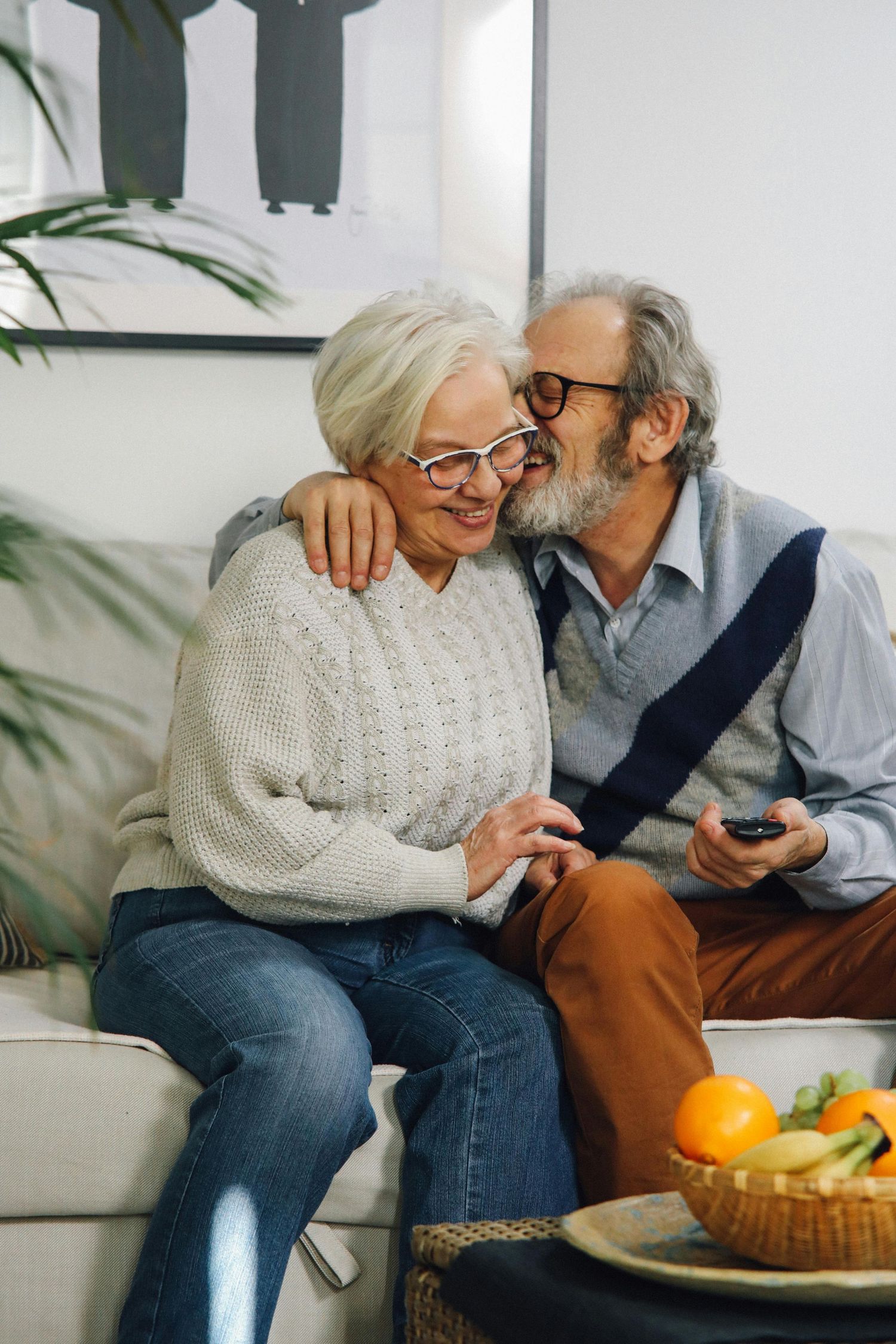Senior couple relaxing together on couch with fresh fruit and digital device, sharing intimate moment at home.