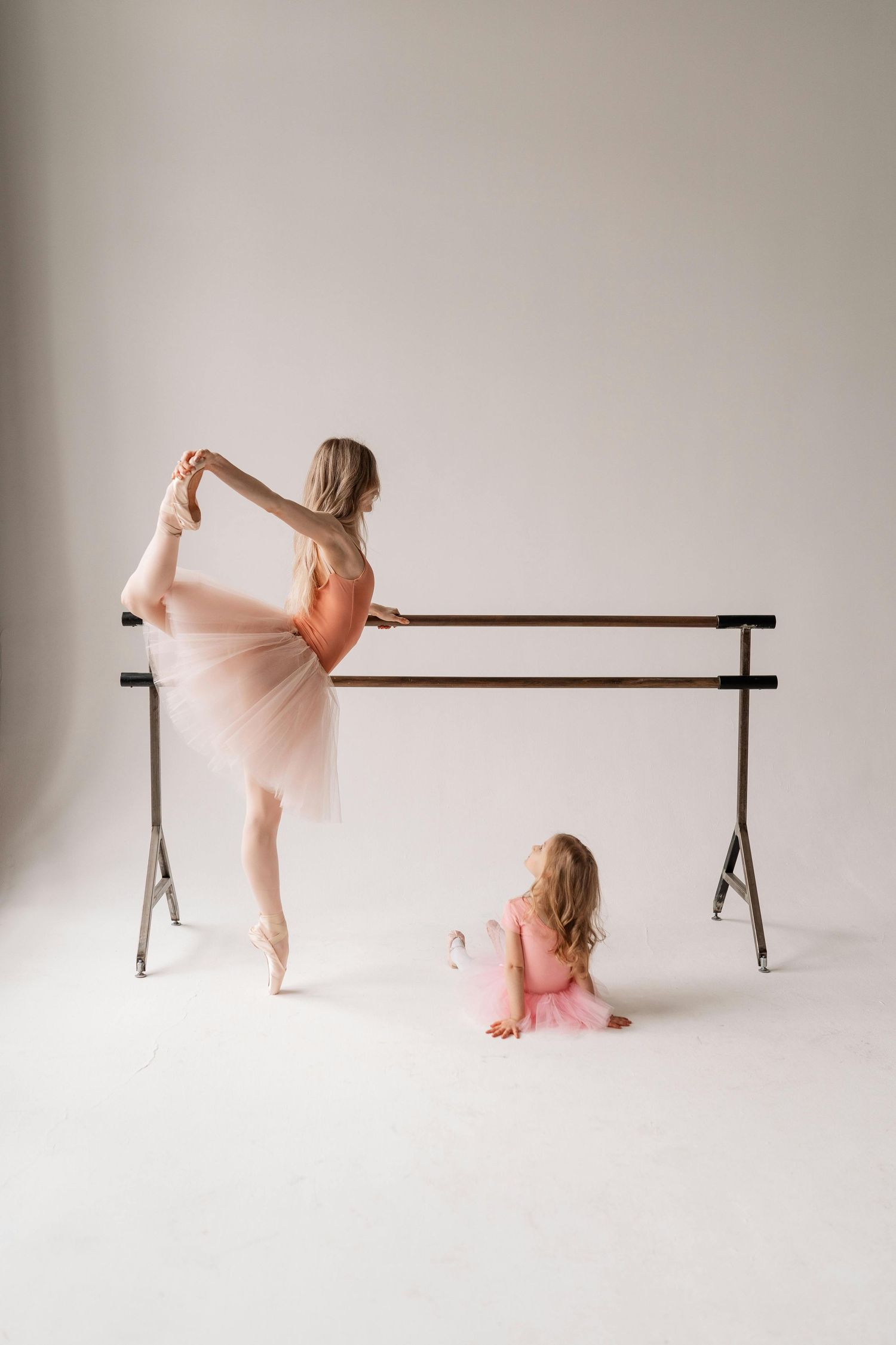 Studio portraits arizona.Two dancers in pink tutus practice ballet at a barre, with one standing en pointe while another sits on the floor.
