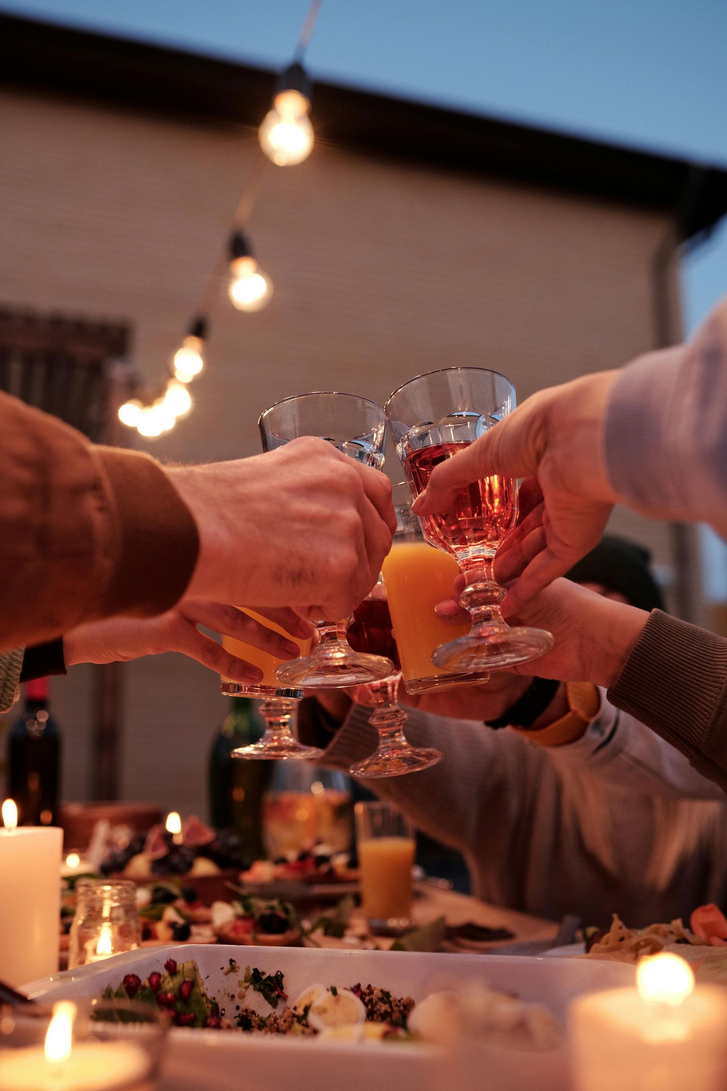 Multiple hands raise wine glasses for a toast at a candlelit dinner table with string lights overhead.
