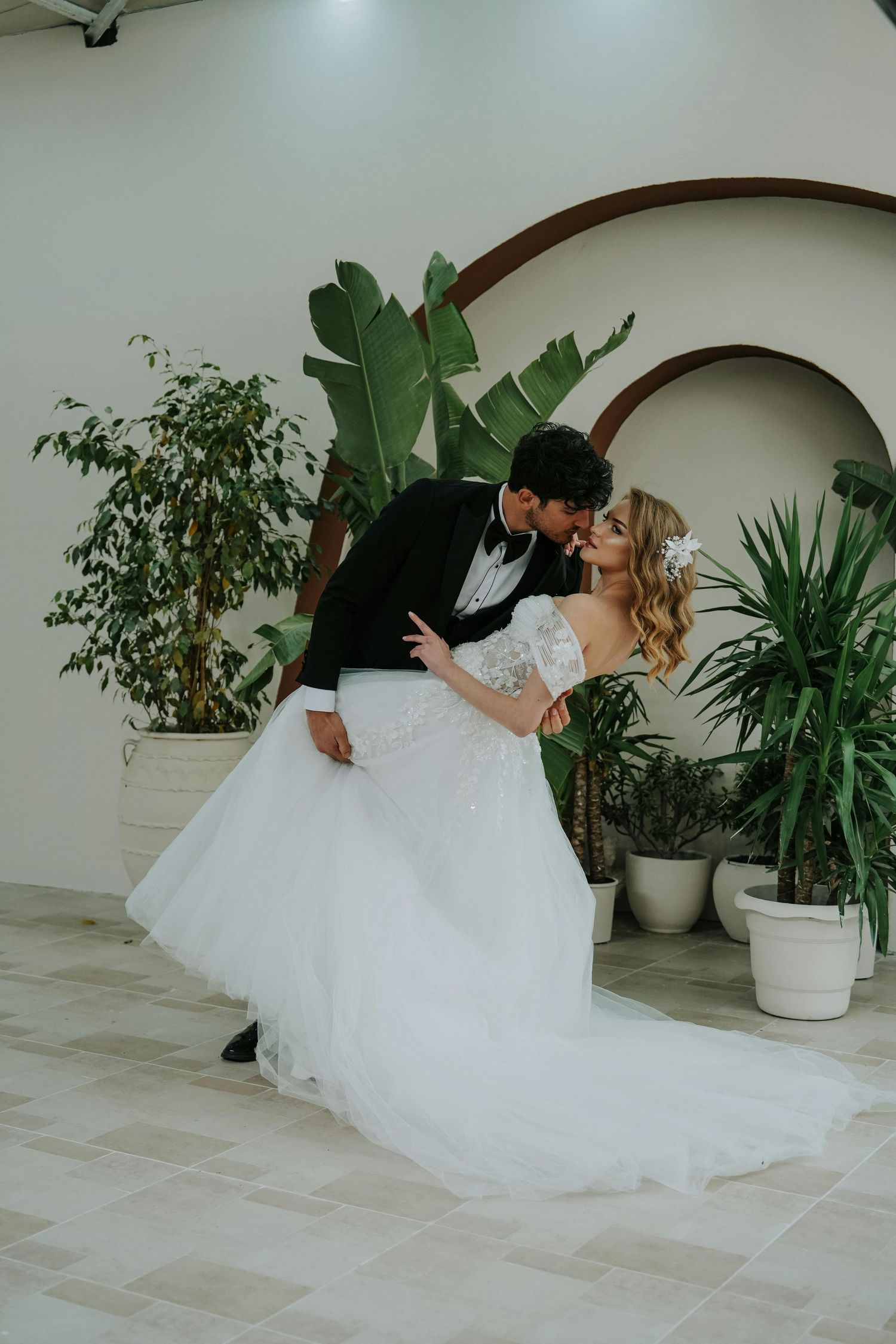 A romantic wedding couple dips and dances together in an indoor space filled with potted green plants and white walls.  Award Winning.