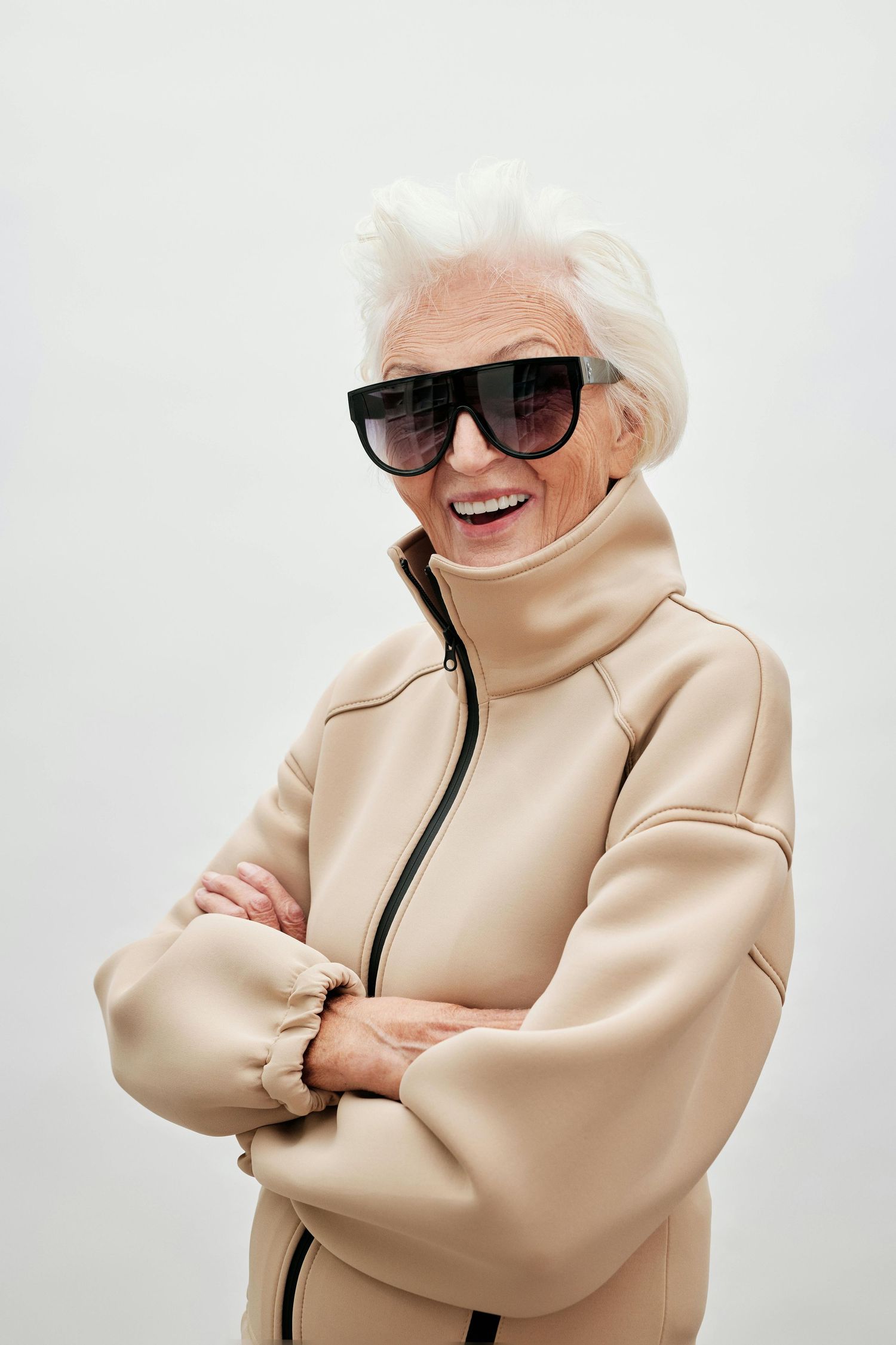 best photographer Arizona. Stylish senior wearing beige turtleneck jacket and designer sunglasses poses with crossed arms and bright smile.