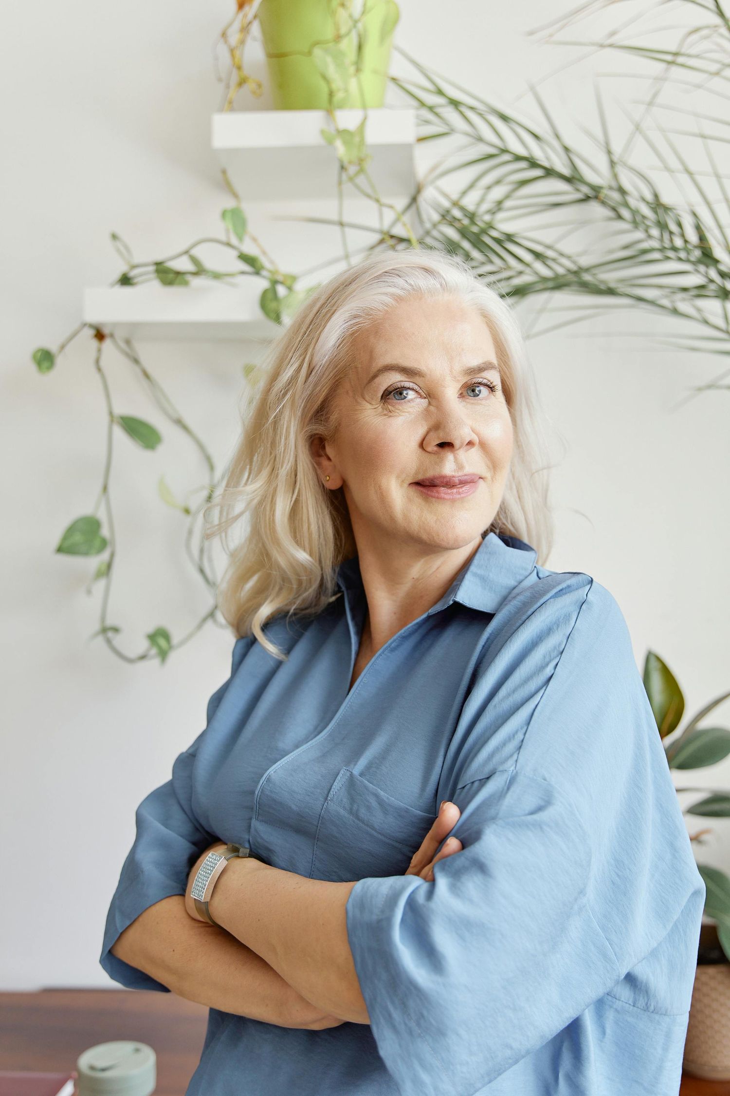 Joyful figure in denim blue shirt poses expressively against white wall with green plants.