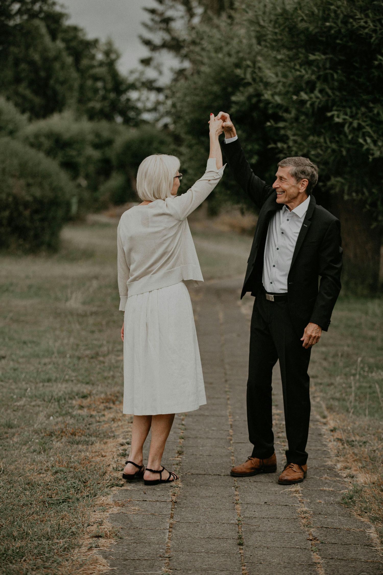 Couple in formal attire dancing and twirling together on a tree-lined pathway in a romantic outdoor setting.