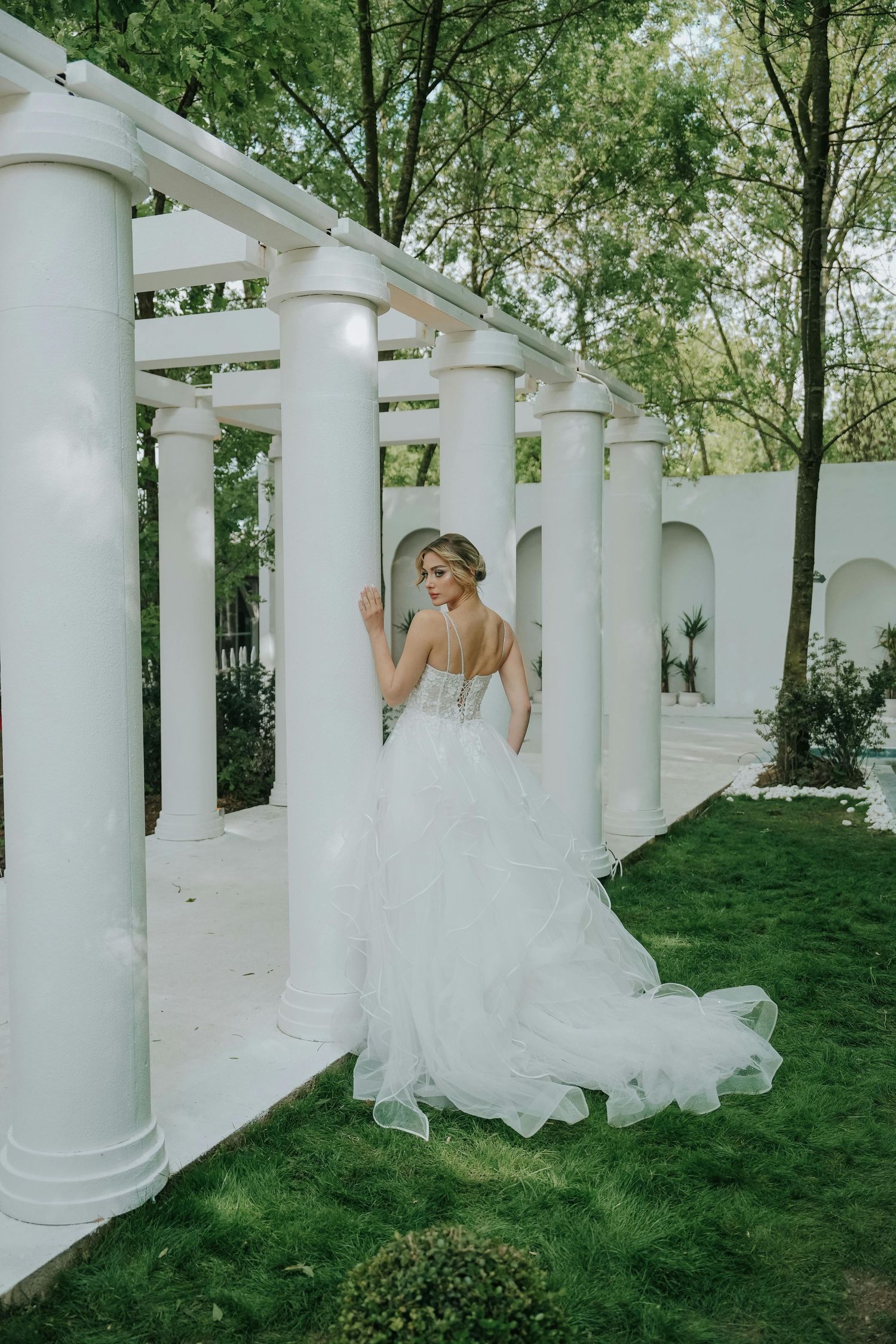 A bride in a flowing white gown poses elegantly among white classical columns in an outdoor garden setting. Leading Arizona Photographer.