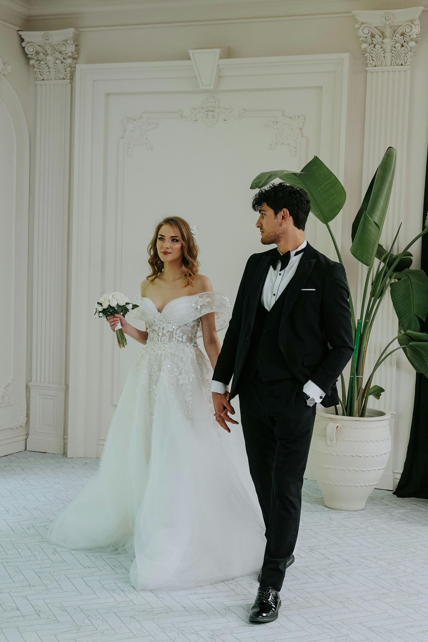 A couple in wedding attire walks together in an elegant white room with ornate molding and potted plants. Top Photographer Scottsdale.