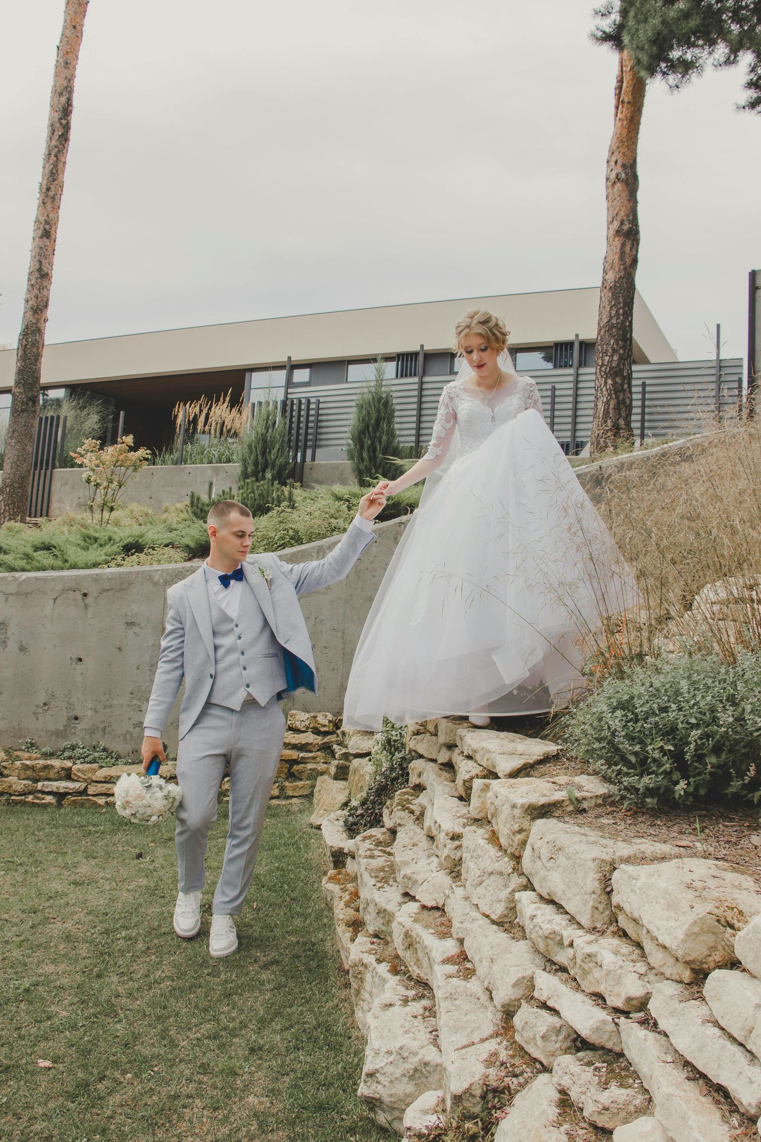 A couple in wedding attire hold hands on a landscaped stone terrace with pine trees and modern architecture in background. Arizona Prescott