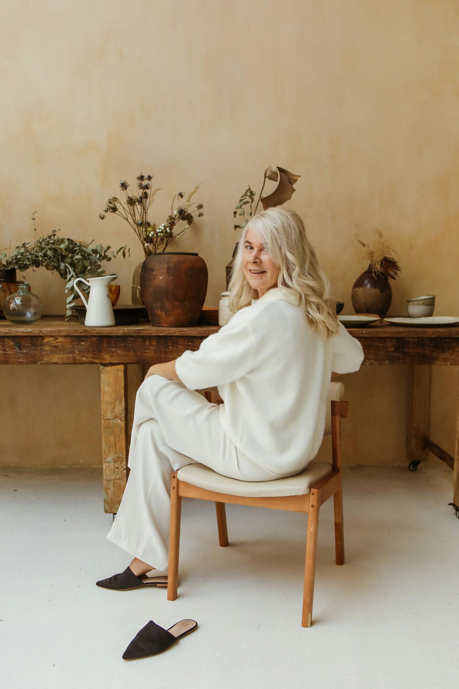 Senior Portraits Flagstaff. A person in a white blanket sits in a wooden chair against a beige wall with rustic shelving displaying pottery.